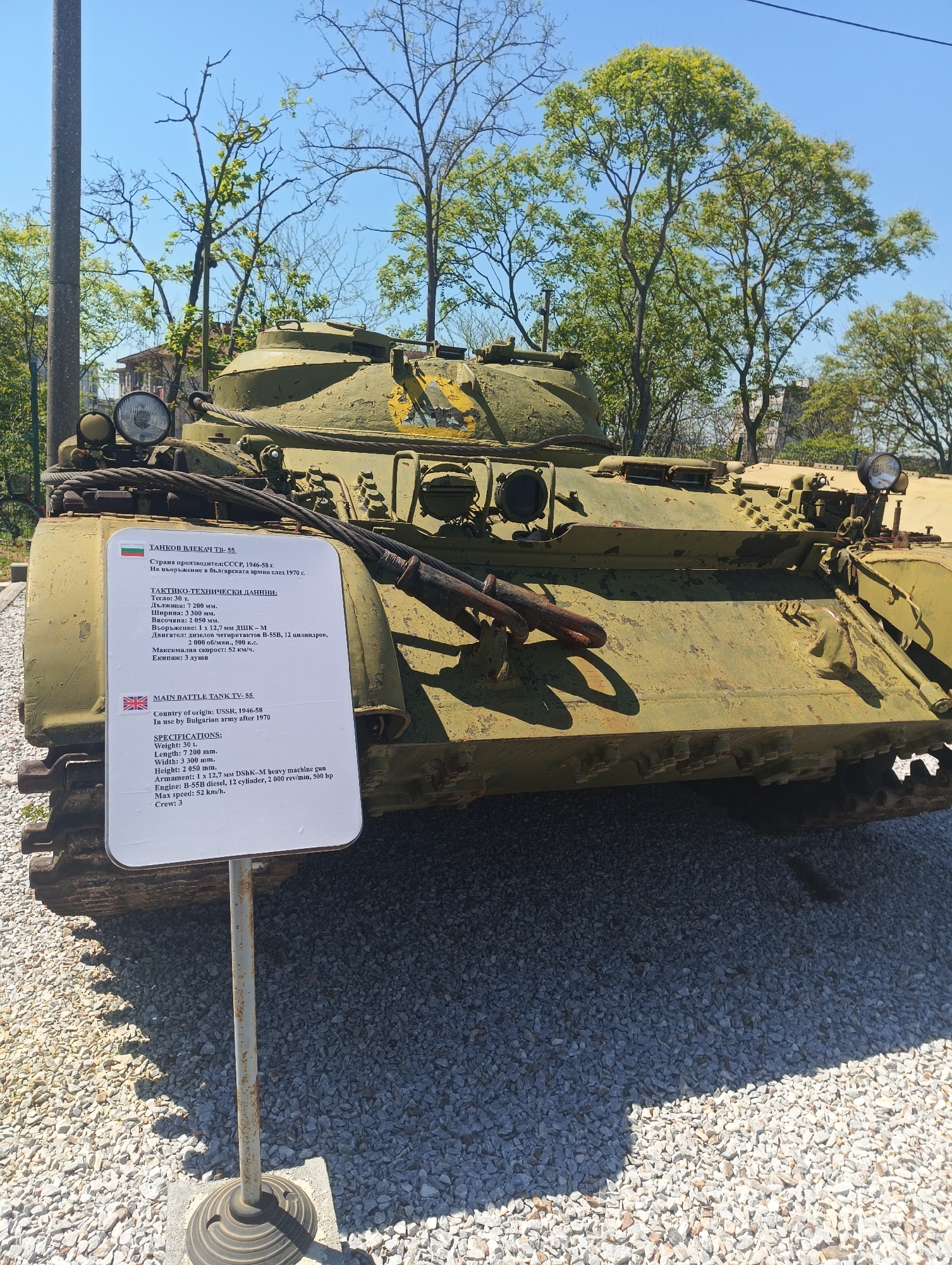 The image depicts a green-painted, presumably decommissioned, TV-55 main battle tank displayed outdoors on a gravel surface. Behind the tank are trees and a partial view of a building. A sign in front of the tank provides information about it in both Bulgarian and English. The sign indicates the tank was manufactured in the USSR between 1946-1958 and was in use by the Bulgarian army after 1970. The specifications listed include a weight of 30 tons, a length of 7200 mm, a width of 3300 mm, a height of 2050 mm, an armament of 1 x 12.7 mm DShK-M heavy machine gun, a B-55B diesel engine with 12 cylinders producing 500 hp, and a maximum speed of 52 km/h. The crew is listed as 3. The location is likely in or near the city of Yambol, Bulgaria, based on the Bulgarian language on the sign.