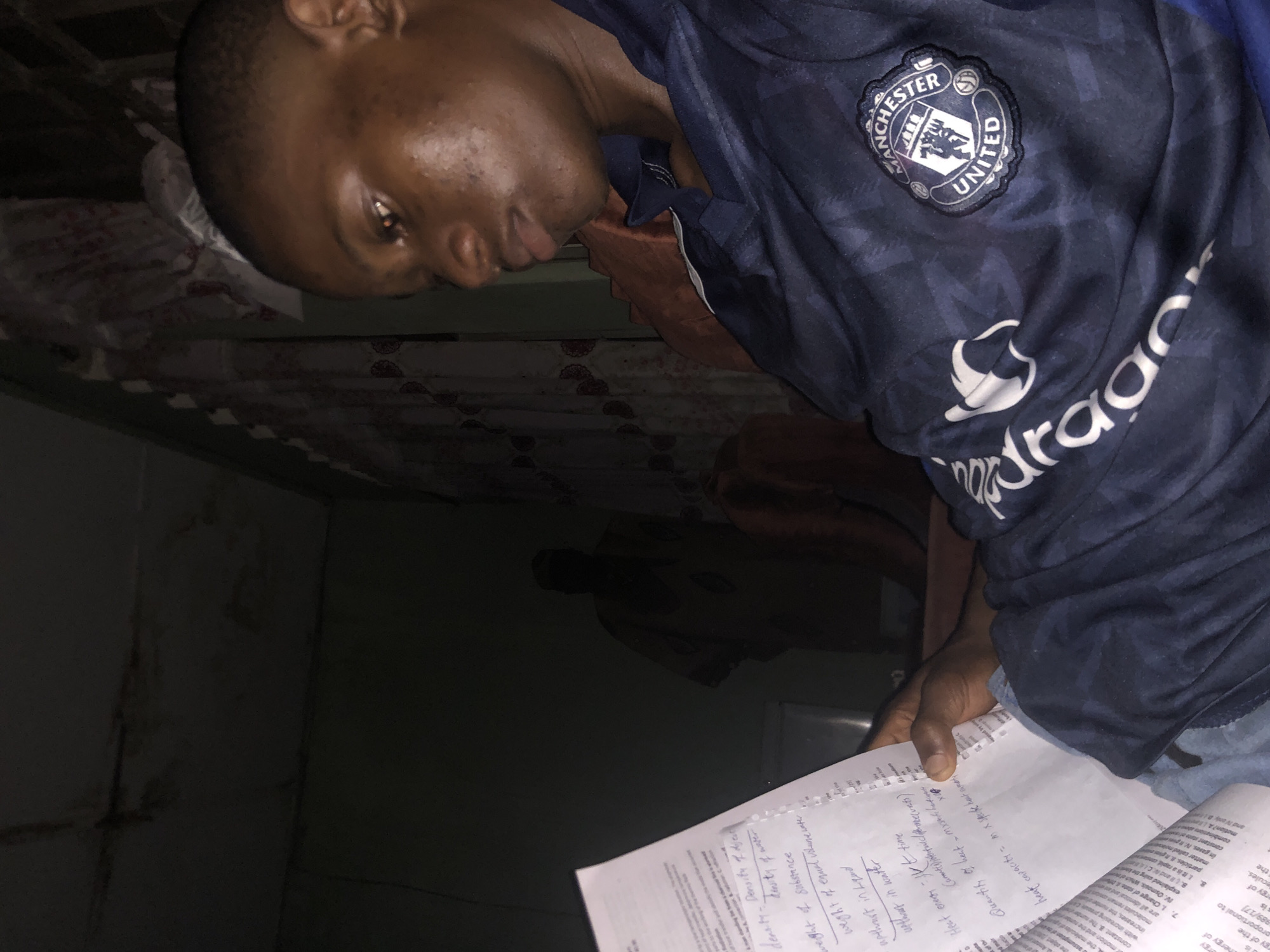 An individual wearing a Manchester United jersey is seated indoors, illuminated by a light source from above. They are holding and reading papers with handwritten notes and printed text. The room is dimly lit, making it difficult to discern further details about the environment. It is not possible to determine if the image was taken in Atani, Nigeria, or confirm the location with the provided information.