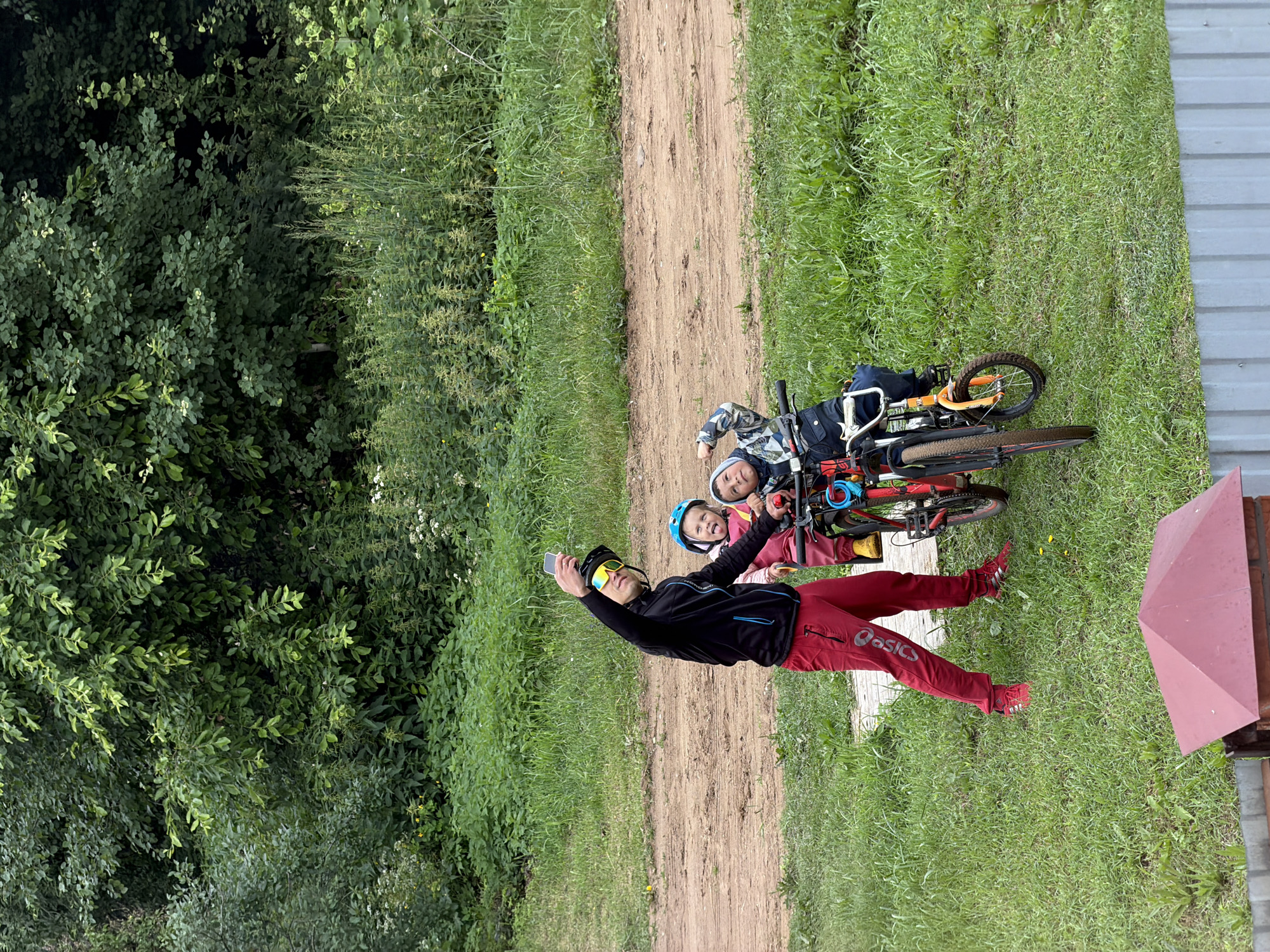 In Chaikovsky, Russia, a man in red pants and a black jacket with blue accents takes a selfie with two children on bicycles. The man wears yellow sunglasses and a black helmet. The child on the left, in a pink jacket and blue helmet, sticks out their tongue. The child on the right, wearing a grey and blue patterned jacket and a hood, looks up. They are on a dirt path surrounded by lush green grass and dense trees. The scene appears to be a casual outing, possibly a park or rural area, given the natural landscape and the presence of bicycles. A red structure with a pointed roof is visible in the foreground, partially obscured by a grey fence.