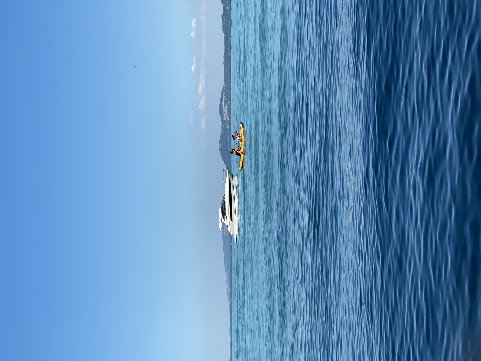 The image shows a body of water, likely a lake, under a clear blue sky. In the middle distance, a white motor yacht is cruising on the water. Closer to the foreground, two people are in a yellow kayak, paddling. The shoreline, with rolling hills and some vegetation, is visible in the background, suggesting an outdoor recreational setting. The scene evokes a sense of calm and leisure.