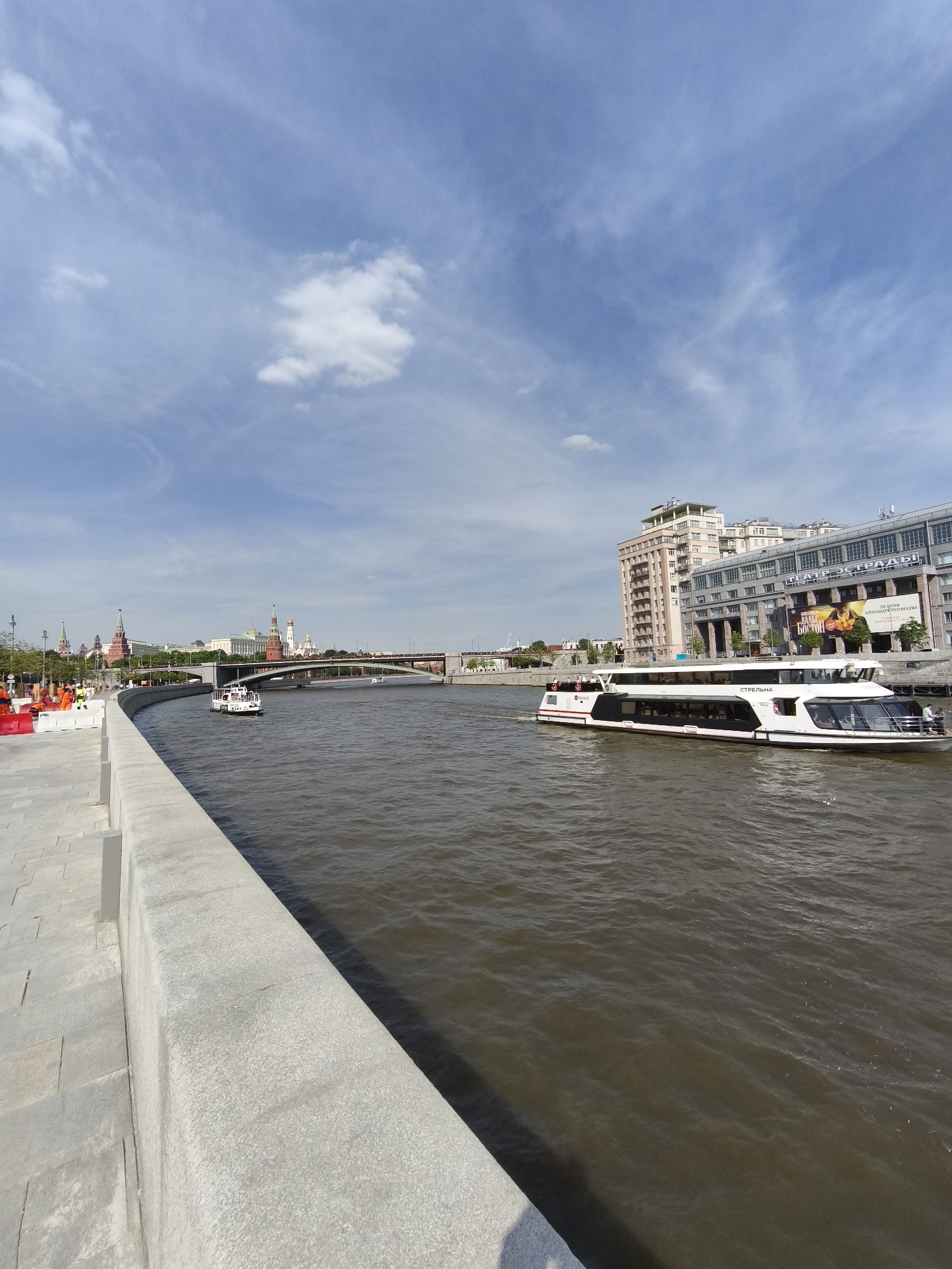 The image displays a panoramic view of the Moskva River in Moscow, Russia, captured on a clear, sunny day with a partly cloudy sky. The perspective is slightly elevated, looking across the river from a concrete embankment on the left.
In the foreground, a light-colored, textured concrete embankment or quay runs along the left side, curving gently to the right as it recedes into the distance. A paved walkway, composed of light gray stone or concrete slabs, extends from the embankment toward the left foreground.
The Moskva River, occupying the central and lower right portions of the frame, exhibits murky, dark brown water with visible ripples and small waves. Two passenger boats are traversing the river. The most prominent boat, on the right side of the river, is a large, modern, two-deck white vessel with black and red accents, named "СТРЕЛЬНА" (Strelna) in Cyrillic script visible on its side. Several individuals are present on the open forward deck of this boat. Further down the river, in the middle-left, a smaller, white, single-deck passenger boat is also in motion.
The far bank of the river showcases significant urban architecture. On the left, a portion of the Moscow Kremlin is distinctly visible, characterized by its red brick walls, numerous towers with green spires, and the golden onion domes of its cathedrals. To the right of the Kremlin, two bridges span the river; the closer bridge is an arched structure.
On the right bank, behind the large "СТРЕЛЬНА" boat, a series of buildings line the river. A tall, light-colored building with multiple stories and windows is visible. Adjacent to it on the right is a large, classical-style building with columns and large windows on its facade. A substantial poster or banner adorns this building, displaying text including "ТЕАТР ЭСТРАДЫ" (Estrada Theatre) in Russian, along with other Cyrillic text and images that appear to be an advertisement.
The sky is a bright, pale blue, featuring scattered thin, white cirrus clouds and a few smaller, fluffier cumulus clouds. Sunlight casts clear shadows, indicating a high sun position. On the near bank, just beyond the concrete embankment to the left, there are signs of ongoing work or temporary installations, including orange safety barriers and faintly visible figures that appear to be construction workers in high-visibility vests.