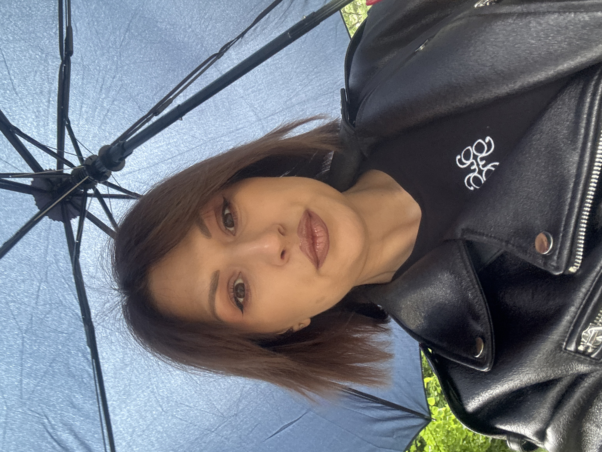 A woman with brown hair is holding a blue umbrella. She is wearing a black shirt with a white logo and a black leather jacket. The location appears to be outdoors, with greenery visible in the background. The photograph was taken from a low angle, looking up at the woman and the umbrella. The setting cannot be confirmed as Sofia, Bulgaria from this image alone.