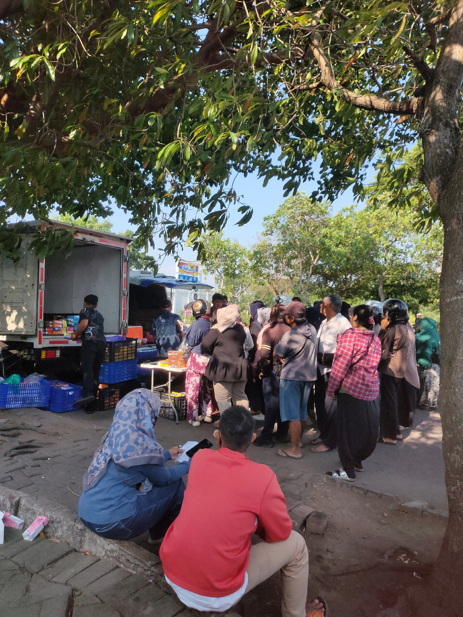 The image depicts an outdoor scene in the city of Ngasem, Indonesia, likely a marketplace or a distribution point. In the background, a white truck with its rear doors open is visible, loaded with goods. Several people are gathered around the truck, some appearing to be interacting with the truck's contents. A table is set up, with crates and boxes arranged on and around it.
In the foreground and mid-ground, a crowd of people is present, dressed in casual attire, with some wearing headscarves. The people are standing and observing, with some engaged in conversations. One person in the foreground, wearing a red shirt and tan pants, is seated. Another person, wearing a blue hijab and jeans, is also seated and looking at a phone.
The environment appears to be a paved area with some dirt and vegetation. Large trees with green foliage are casting shadows over the scene, indicating it is daytime. The sky is clear and blue. The overall impression is one of a busy, communal activity, possibly related to the distribution or sale of goods from the truck.