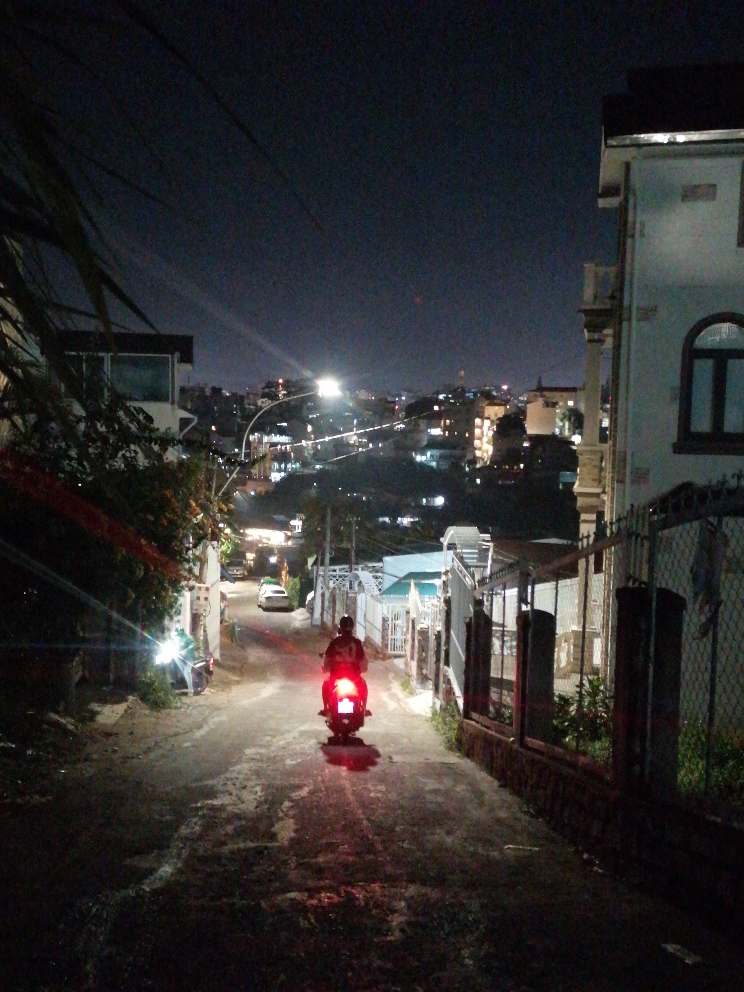 The image depicts a nighttime view in Da Lat, Vietnam, looking down a narrow, paved street towards a distant cityscape. A person is riding a scooter away from the viewer, the tail light glowing red. On the left side of the road is a building with a visible light, foliage, and parked scooters. On the right, a fence runs along a property. Streetlights illuminate the distant city on a hill. The sky is dark.