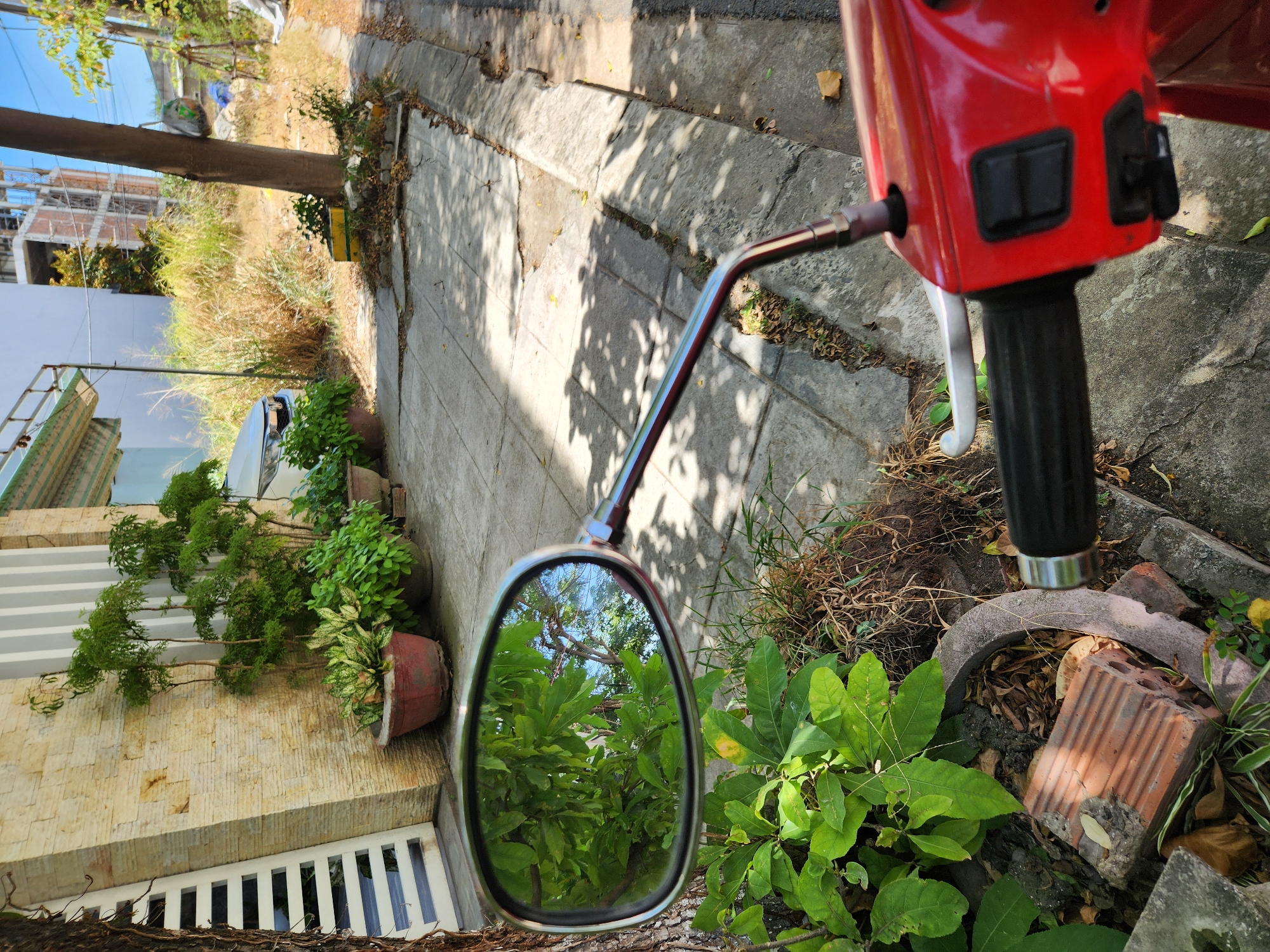 This image captures a street-level view in Đà Nẵng, Vietnam, likely in a residential or semi-commercial area. The foreground is dominated by the handlebars and a side mirror of a red scooter. The mirror reflects lush green foliage and a bright blue sky, suggesting it's a sunny day. The scooter is parked on a paved walkway, bordering a street with a concrete curb. To the left of the scooter, a beige-toned wall with vertical white accents is visible, alongside several potted plants. Further in the background, a white car is parked, and beyond that, a patch of overgrown vegetation leads to a partially constructed building with scaffolding, indicative of ongoing development. The overall impression is that of a typical urban scene in Vietnam, with a mix of residential elements and construction.