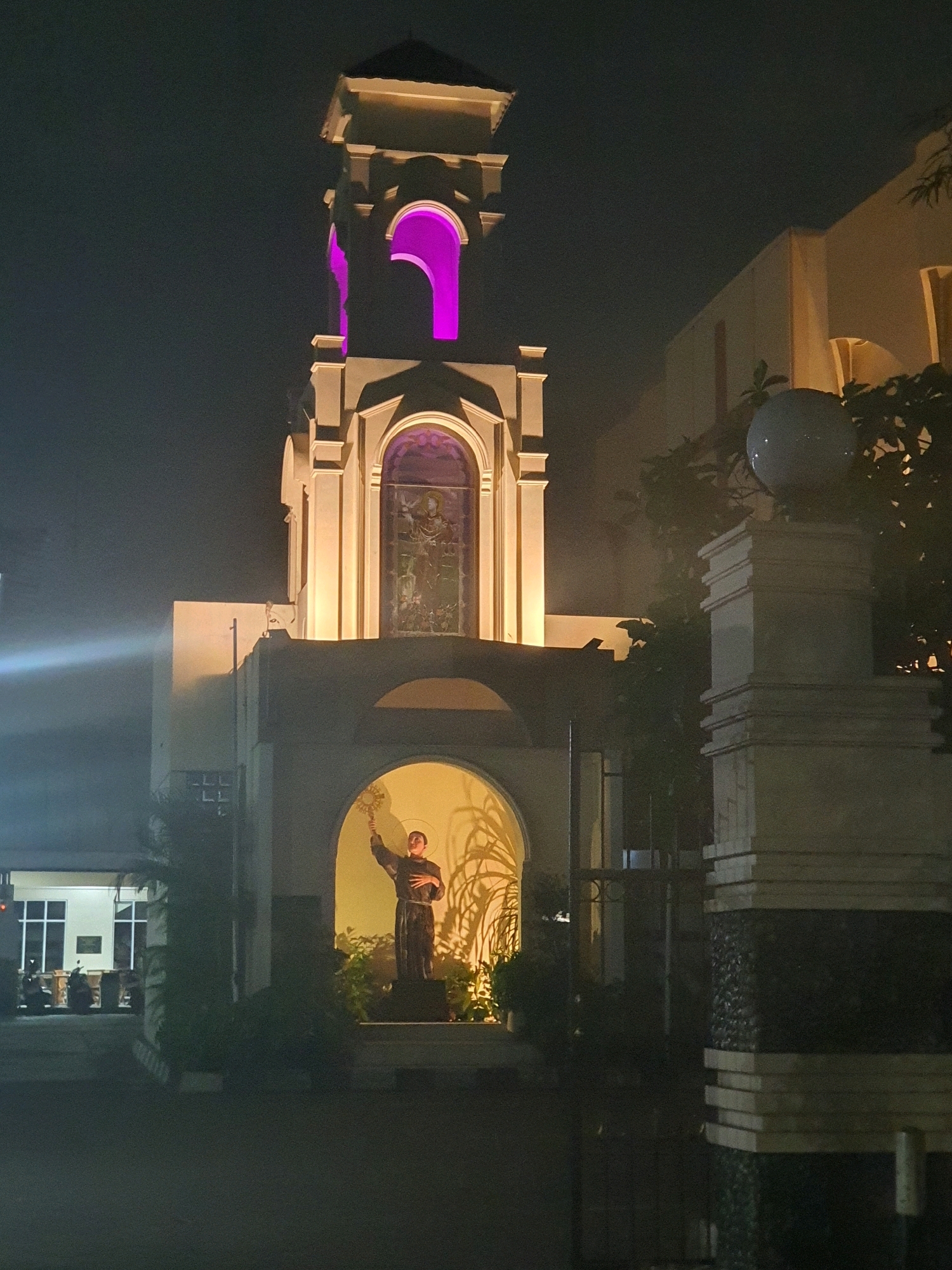 At night, a statue of a robed figure holding a religious object is illuminated within an arched alcove of a building in Central Jakarta, Indonesia. The statue is surrounded by lush green plants, creating a dramatic contrast against the warm yellow light emanating from behind it. Above the alcove, a tower with arched openings glows with vibrant purple light. To the right of the building, a large, spherical light fixture sits atop a stone pillar. In the background, a modern building with large windows and parked motorcycles is visible. The scene is captured from a low angle, with the dark foreground suggesting a street or pathway.