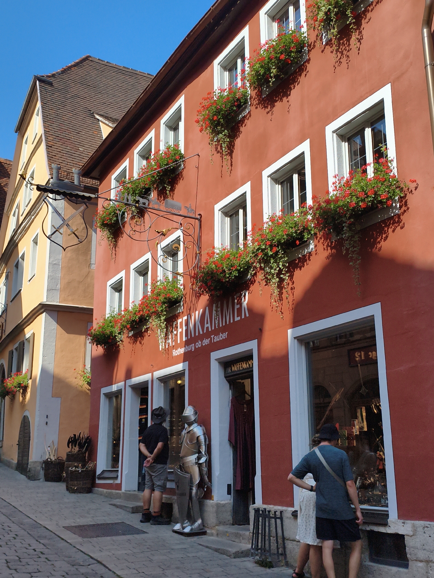 The image captures a street scene in Rothenburg ob der Tauber, Germany, with a prominent red building dominating the right side of the frame. The building features multiple windows adorned with red flowers in window boxes, creating a vibrant display against the red facade. A sign above one of the windows reads "WAFFENKAMMER Rothenburg ob der Tauber," indicating a weapons chamber or shop. In front of the shop, a full-sized suit of armor stands as a decorative display. Two people are visible near the entrance of the shop: a man wearing a hat and shorts, and another person, partially obscured, wearing a dress and a hat. The street is paved with cobblestones. To the left of the red building, a lighter-colored building with a brown tiled roof is partially visible. Several barrels are placed along the side of the street, and some vegetation is visible in the background. The sky is clear and blue.