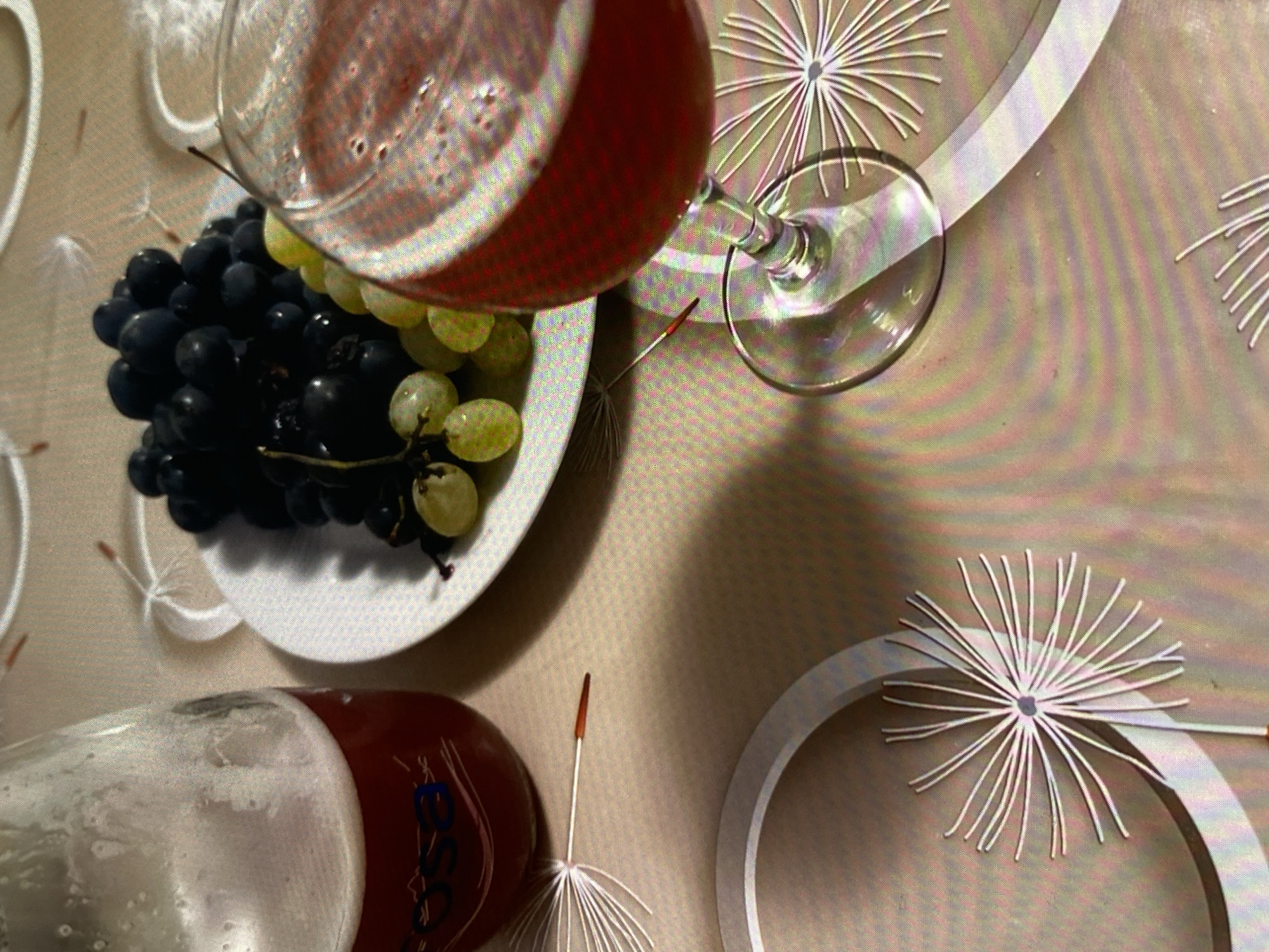A still life arrangement features a glass of red liquid and a plate of grapes on a patterned tablecloth. To the left, a glass bottle with a red liquid and foam is partially visible. The bottle has blue lettering. The plate contains a bunch of dark purple grapes and a smaller bunch of green grapes. Several dandelion seed heads are scattered on the tablecloth, some intact and some broken. The tablecloth itself has a light brown base color with a repeating pattern of concentric circles and radiating lines that resemble dandelion seed heads. The scene is lit from above, casting shadows on the tablecloth. The image is a close-up, angled shot focusing on the food and drink items.
