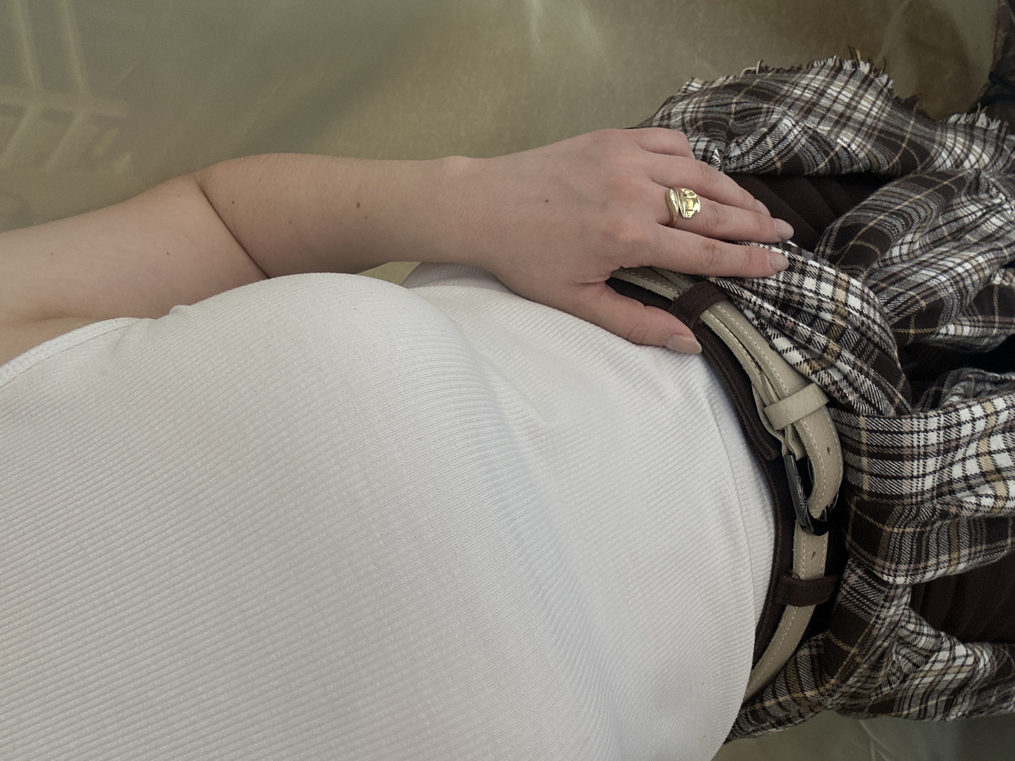A top-down view shows a person lying down, wearing a white ribbed tank top. The person's left arm is bent with their hand resting on a brown plaid garment with a tan belt. A gold ring is visible on the person's hand. The background is a beige surface.