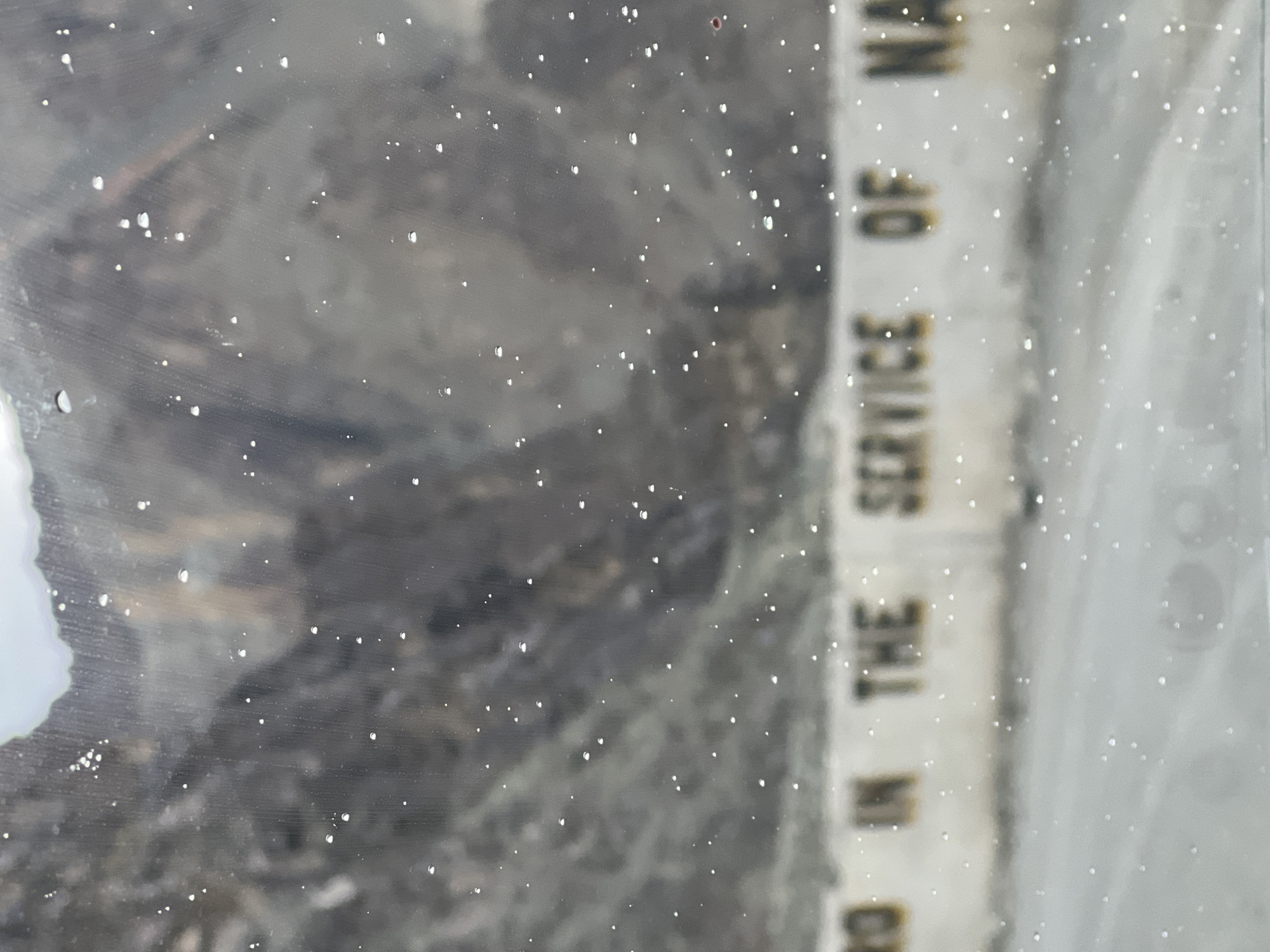 The image shows a blurred view of a mountainous landscape through a surface with numerous water droplets and small white specks. In the foreground, a low, white wall with yellow lettering is visible. The visible text on the wall reads, "IN THE SERVICE OF NA". The wall appears to be a guardrail or retaining wall, likely along a road. The background consists of blurred, rocky mountains with patches of light and shadow. The overall impression is of a scene viewed from inside a vehicle on a wet or misty day, possibly in a mountainous region. The text suggests a patriotic or service-oriented message, common on infrastructure in certain locations. The location in Khaltse, India, would imply a mountainous terrain, possibly along a road like the Leh-Srinagar highway.