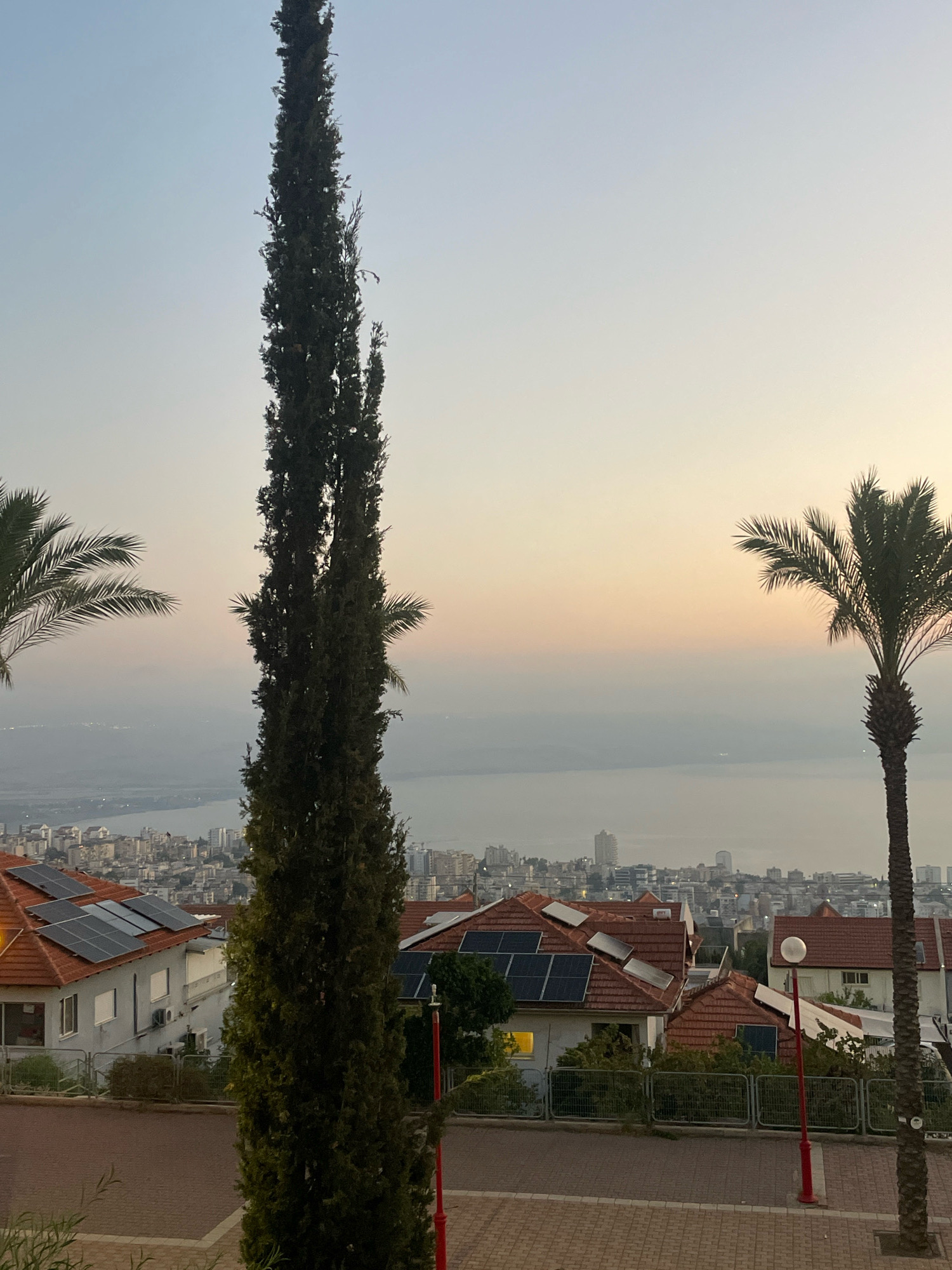 This image depicts a scenic view of Tiberias, Israel, at either sunrise or sunset. In the foreground, a tall, slender cypress tree dominates the left side of the frame, its dark green foliage contrasting with the pale blue and pink hues of the sky. To the left and right of the cypress tree, palm fronds are visible, suggesting a Mediterranean climate.
Below the trees, a residential area with red-tiled roofs is seen. Several houses are equipped with solar panels, indicating a modern and environmentally conscious community. The houses are nestled on a hillside, offering a panoramic view of Tiberias. In the midground, the city sprawls out, with buildings of varying heights and architectural styles. Beyond the city, a body of water, likely the Sea of Galilee, stretches across the horizon, its surface appearing calm and slightly hazy. The distant cityscape is softened by the atmospheric conditions of either dawn or dusk, with soft light illuminating the scene. The overall impression is one of tranquility and natural beauty, blended with urban development.