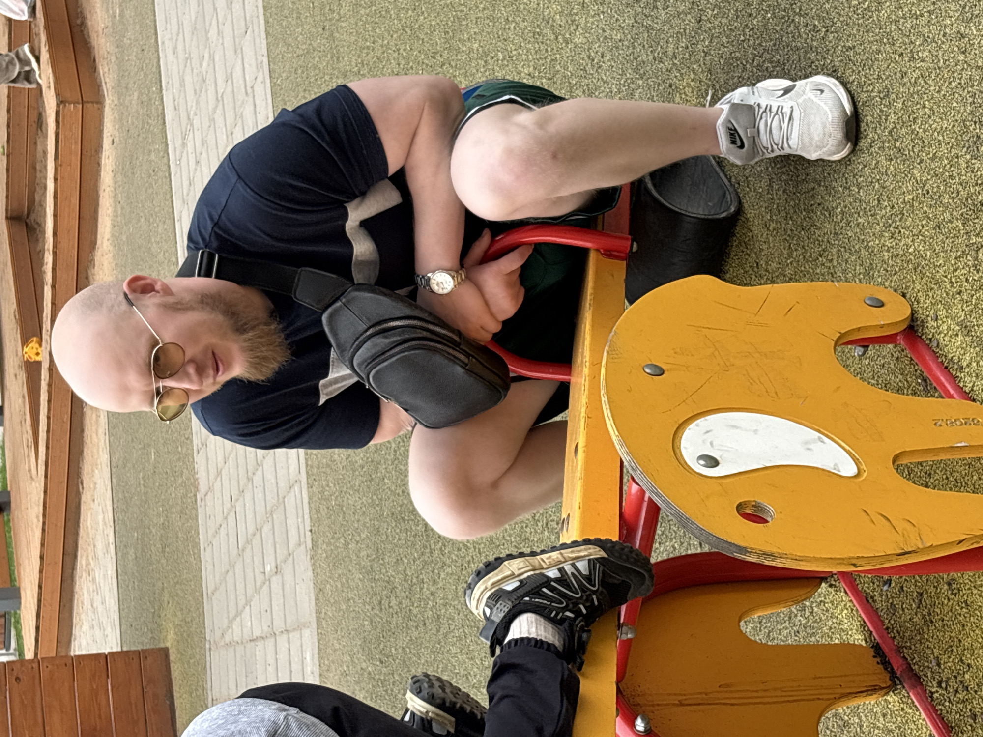 A bald, bearded man wearing a dark blue shirt, light-colored sunglasses, a watch, and a black fanny pack is seated on a yellow and red children's playground seesaw. He has one leg bent, with a white Nike sneaker visible, and the other leg extended to the other side of the seesaw, resting his foot, which is wearing a black sneaker, on the other seat. The background shows a park with a playground surface and brick pathways. The photograph may have been taken in Izhevsk, Russia, as requested, but there is no definitive, verifiable information to confirm this.