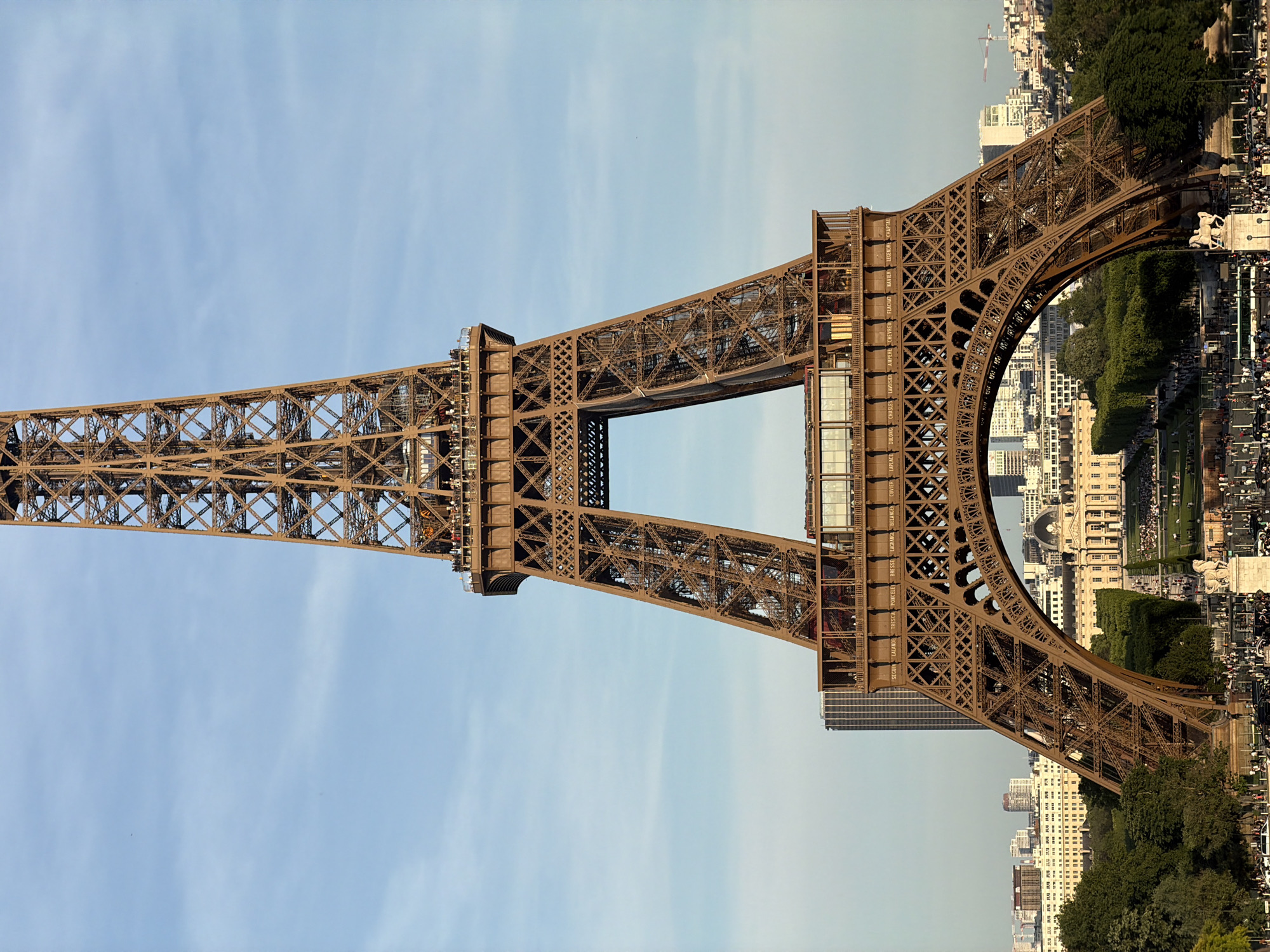 The image shows the Eiffel Tower in Paris, France. The tower is made of brown iron and features an intricate lattice structure. The sky is a pale blue with some wispy clouds. The base of the tower is surrounded by trees, buildings, and people. The buildings appear to be a mix of residential and commercial. The people are likely tourists visiting the iconic landmark.