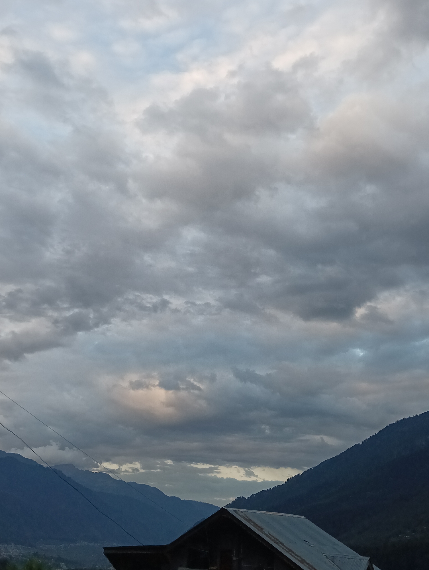 The image shows a cloudy sky over a mountainous landscape with a building in the foreground. The sky is filled with a mix of gray and white clouds, with some patches of blue visible. The mountains are dark blue and covered in trees. A portion of a building with a tin roof is visible at the bottom of the image. The scene appears to be in Vashisht, India, a town known for its natural beauty and hot springs, located in the Kullu Valley of Himachal Pradesh. The overall mood of the image is serene and slightly dramatic due to the cloudy sky.