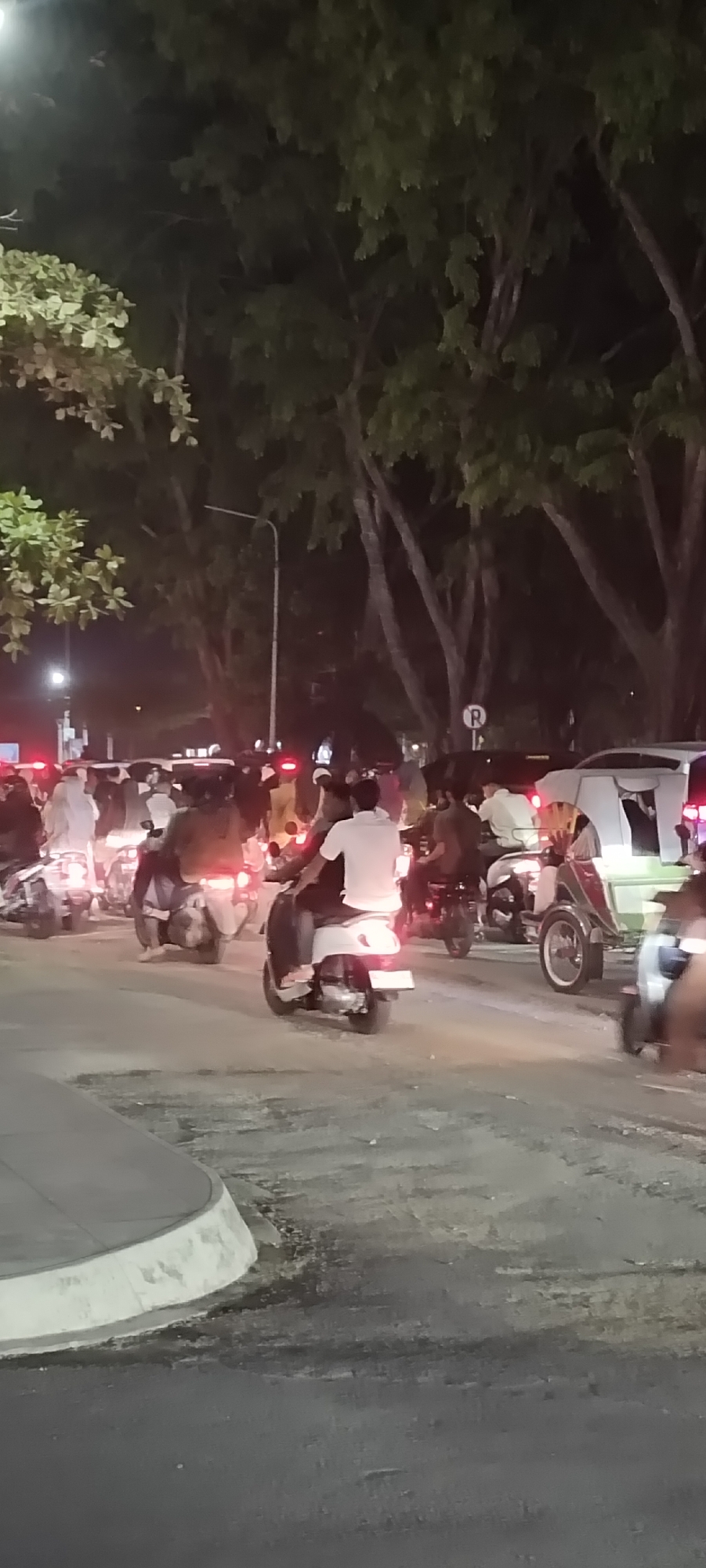 The photograph, taken at night, shows a traffic jam in Lhokseumawe, Indonesia. The road is filled with motorbikes, with the backs of several riders visible in the foreground. A vehicle with a sidecar is also present, appearing green and white. The riders are in street clothes, t-shirts and the like. A traffic light can be made out in the background. The sky is dark, and trees line the street, illuminated by street lights. The ground is covered in asphalt.