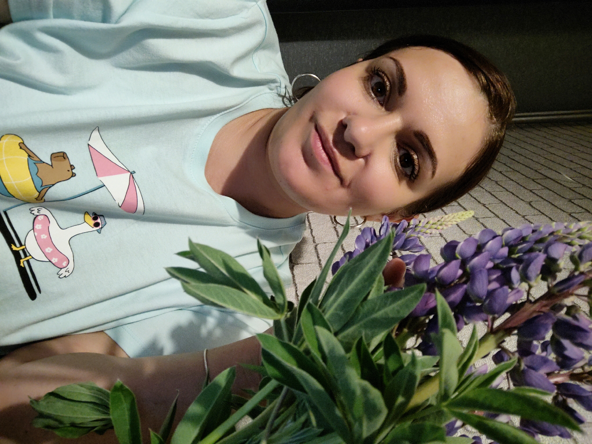 A selfie captured from above shows a woman with light skin, brown eyes, and dark brown hair pulled back, posing with a bouquet of purple lupine flowers. She is wearing hoop earrings and a light blue t-shirt with cartoon images of animals in swimwear. The background appears to be an indoor tiled surface. The location is Марковский, Russia.