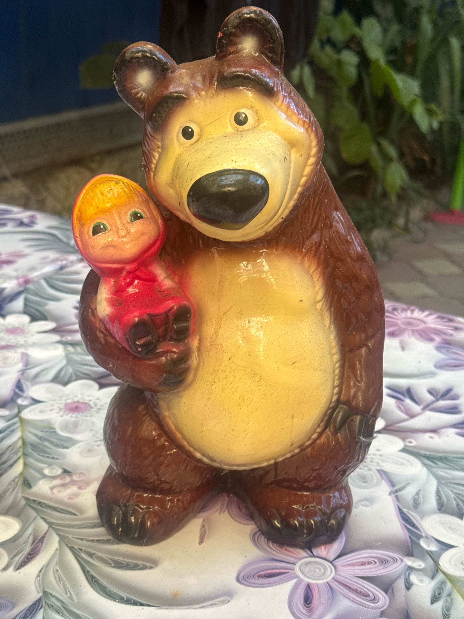 A close-up, eye-level shot captures a brown, plastic figurine of a bear holding a smaller, doll-like figurine. The bear has a yellow face with black eyes and nose, and its ears are rounded and brown. The bear's body is a glossy brown, and its belly is a pale yellow. The doll, with orange hair and rosy cheeks, wears a red dress. Both figurines appear worn, with visible scuff marks and chipped paint. They are positioned on a patterned tablecloth with floral designs in shades of white and purple. The background is out of focus, showing greenery and a dark surface, suggesting an outdoor or semi-outdoor setting. The image is lit from above, casting a slight glare on the figurines. The scene is devoid of any discernible Anapa landmarks.