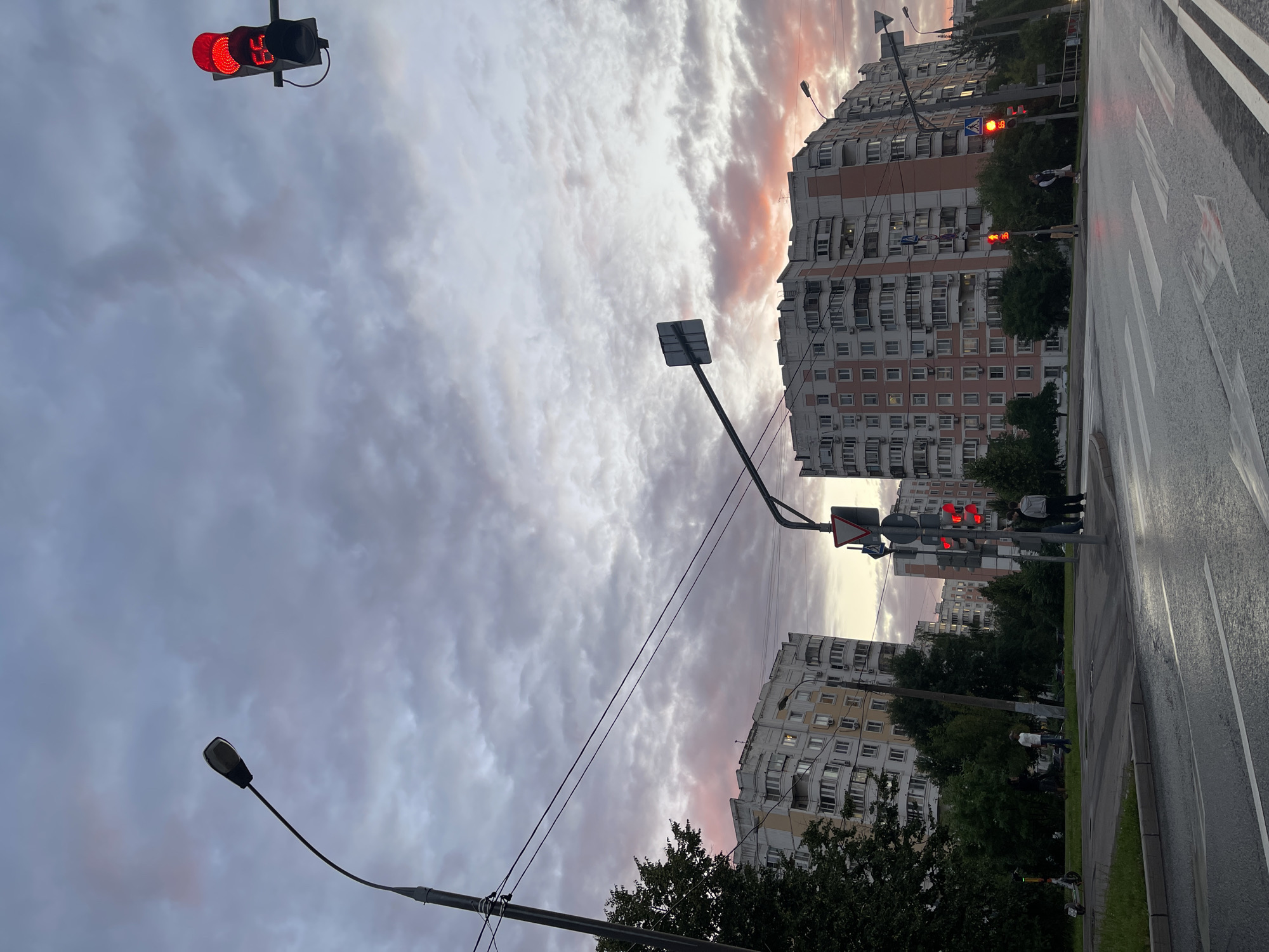 This image depicts a city intersection at dusk, with apartment buildings and traffic signals visible. The sky is filled with dramatic clouds, tinged with pink and purple hues from the setting sun. Several traffic lights are red, including one with a countdown timer displaying "65". People are seen standing on the sidewalk, waiting to cross the street, and an electric scooter is parked on the side. The wet asphalt of the road suggests recent rain. The architectural style of the buildings, characterized by their uniform facades and numerous balconies, is typical of Soviet-era residential blocks found in Moscow, Russia. The presence of streetlights and road markings further confirms an urban environment.