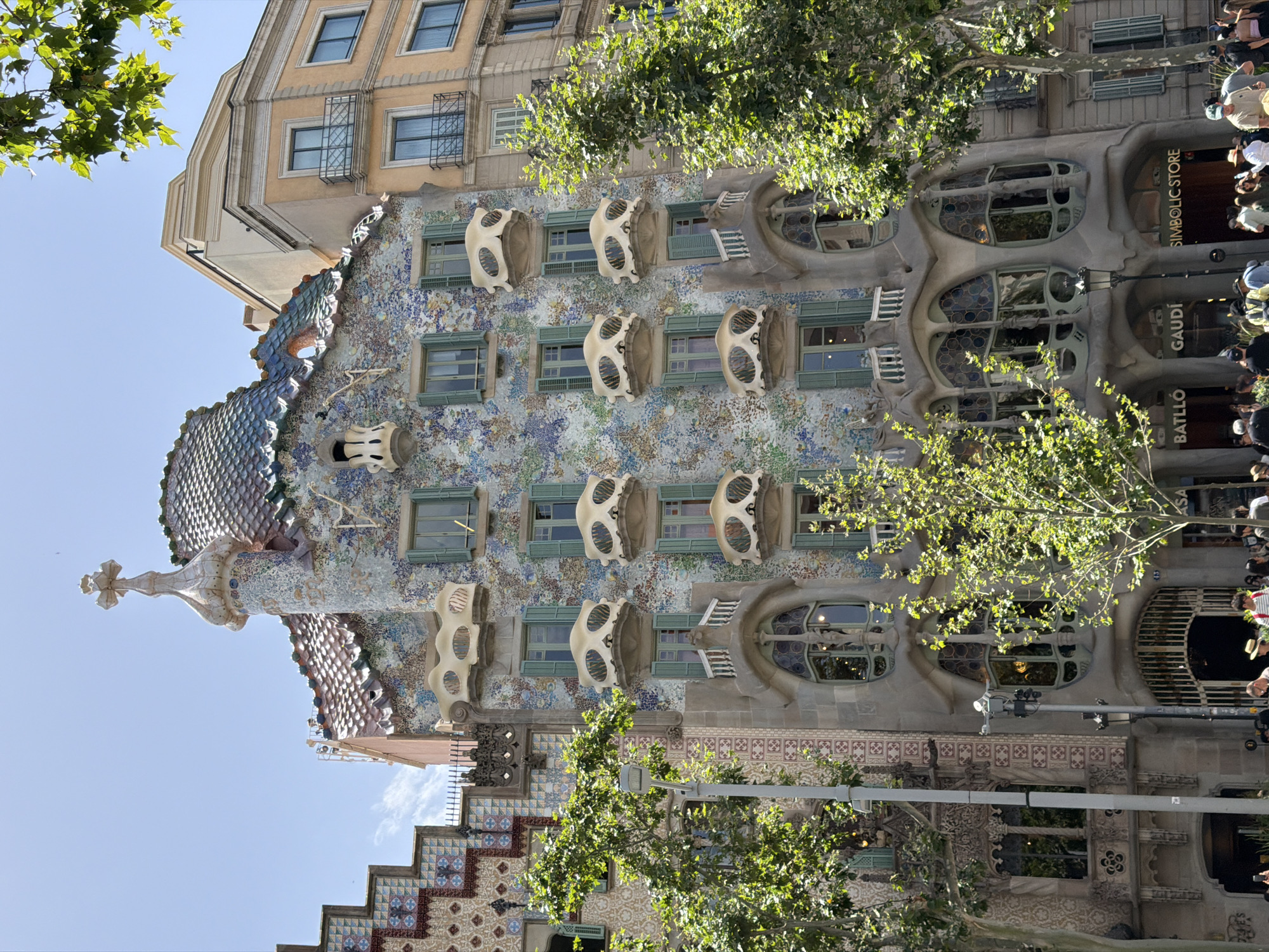 This image captures a street-level view of Casa Batlló in Barcelona, Spain, an iconic building designed by Antoni Gaudí. The facade is adorned with colorful mosaic tiles and features whimsical, organic architectural elements, including bone-shaped balconies and a dragon-scale-like roof. Lush green trees partially obscure the building, casting dappled sunlight on its intricate surfaces.
The scene appears to be a bright, sunny day, likely late morning or early afternoon, given the strong sunlight and clear sky. A crowd of people is visible, gathered on the sidewalk in front of the building, suggesting it is a popular tourist attraction. Some individuals are looking up at the architecture, while others are walking by.
Visible text includes "BATLLÓ," "GAUDÍ," and "SYMBOLIC STORE" on signage near the entrance. The overall atmosphere is one of vibrant urban life and architectural wonder. The perspective is tilted, giving a dynamic and slightly disorienting view of the building and its surroundings. The bright colors of the building and the greenery create a lively and engaging visual experience.
