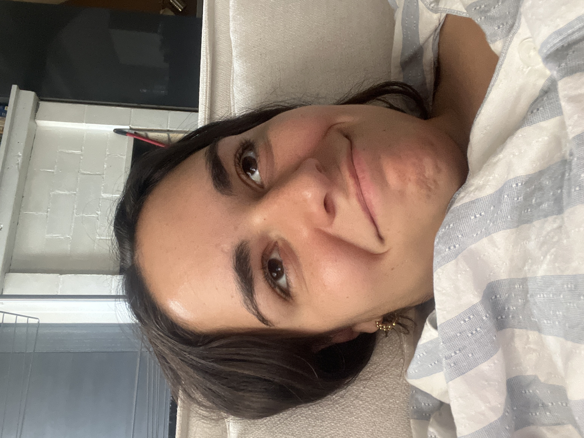 A close-up, high-angle, indoor shot shows the upper body of a young woman lying down, covered by a light blue and white striped comforter. Her dark hair is spread out around her head, and she is looking upwards with a slight smile. Her right ear, adorned with a gold star-shaped earring, is visible. The background features a brick wall painted white and a dark rectangular object, possibly a television screen. The scene is devoid of any discernible Parisian landmarks.