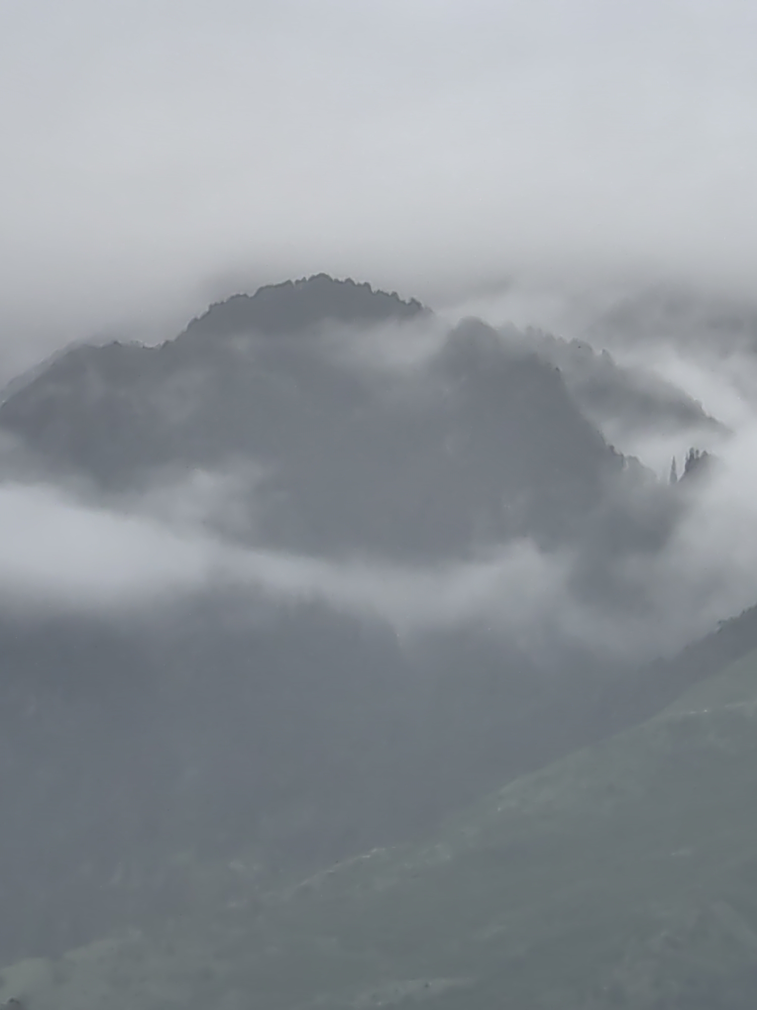 The image depicts a mountainous landscape shrouded in dense fog. The terrain is characterized by rolling hills and peaks, with varying degrees of visibility due to the atmospheric conditions. The foreground shows the slopes of a lower hill, partially obscured by fog. In the mid-ground, a larger mountain range emerges from the mist, its silhouette sharply defined against the hazy sky. The upper parts of the mountains are almost entirely concealed by thick clouds, creating a dramatic and ethereal effect. The color palette is predominantly grayscale, with shades of gray, white, and dark gray dominating the scene. No people or specific man-made objects are clearly discernible in the image, suggesting a natural and remote setting. The overall impression is one of a serene yet mysterious environment, typical of mountainous regions experiencing fog or cloud cover. The location is Vashisht, India.