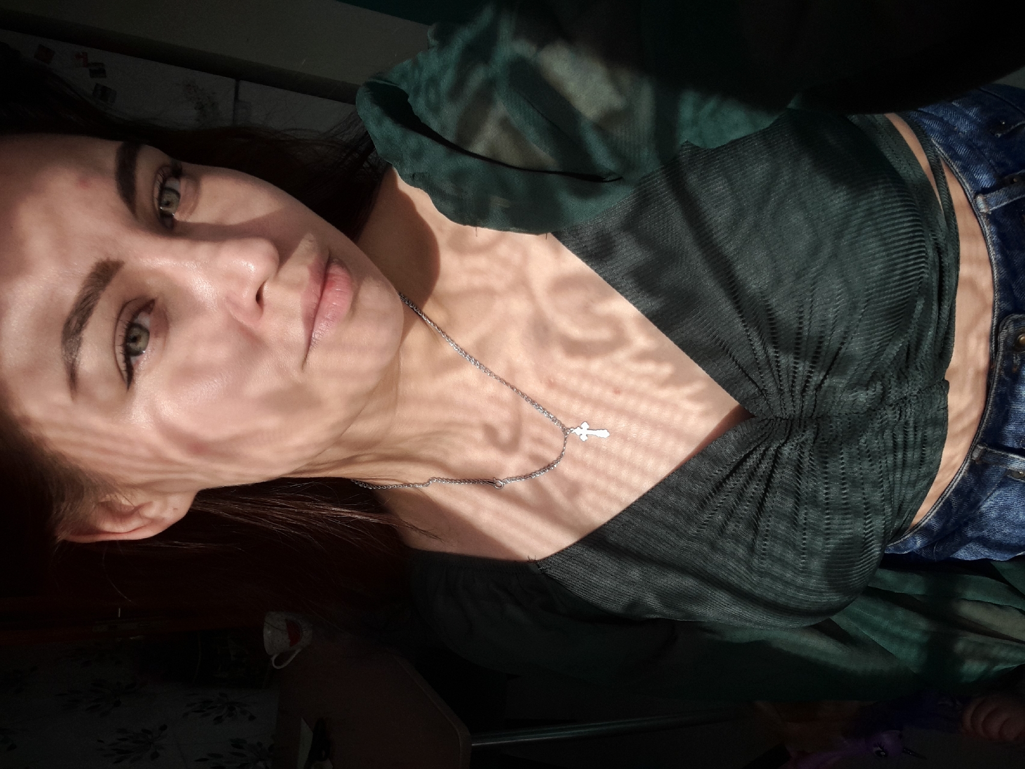 A woman is pictured in a close-up, angled shot. Her face is partially covered by shadows cast by what appears to be a window covering. She has fair skin, light-colored eyes, and dark eyebrows. She wears a dark green, semi-transparent top with a V-neck and a silver necklace with a cross pendant. The neckline of her blue denim jeans is visible. The setting is indoors with a dark background. Location is Unknown, Unknown.