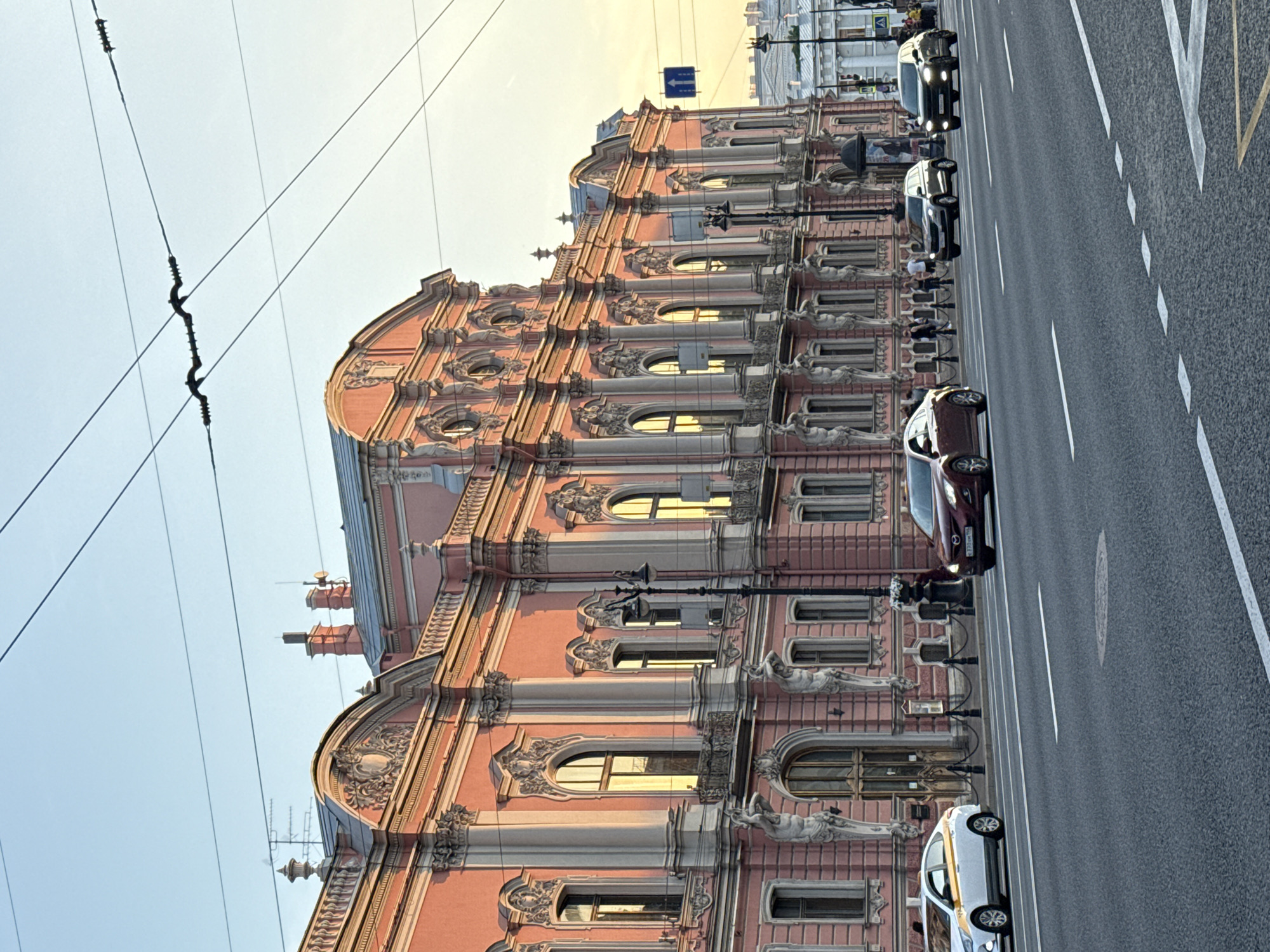 This media file captures a street scene in Saint Petersburg, Russia, with a focus on a grand, ornate pink building. The building features multiple stories with arched windows, decorative sculptures, and intricate architectural details, indicative of classic European design. Overhead power lines crisscross the sky, suggesting an urban environment with a public transportation system.
The street is paved and marked with white lane lines, and several cars are visible, including a dark sedan and a white taxi. The cars are driving along the street, indicating ongoing traffic. People are present, though they appear as small figures in the background, suggesting a normal day in the city.
The lighting suggests it is either early morning or late afternoon, with a warm glow illuminating the building and the sky. The sky is clear and bright, indicating fair weather. The overall impression is one of a historic and vibrant city, with the architecture of the building being a prominent subject. No visible text is discernable in the image.