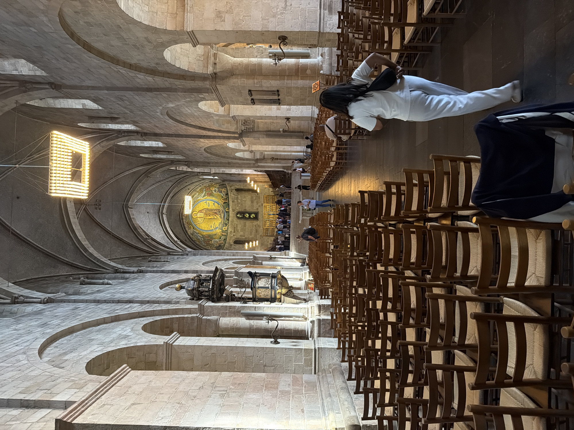 The image shows the interior of Lund Cathedral in Lund, Sweden. The camera is positioned looking down the nave of the cathedral towards the apse. Rows of empty wooden chairs with woven seats fill the floor. The cathedral's architecture is characterized by stone walls, arched ceilings, and large pillars. A woman in white pants and a white t-shirt with a black crossbody bag is walking away from the camera down the center aisle. In the background, there are a few people gathered near the altar area, which is adorned with a large mosaic. A pulpit is visible on the left side of the nave. The lighting appears to be a mix of natural light coming from high windows and artificial light from a large, modern chandelier and wall sconces.