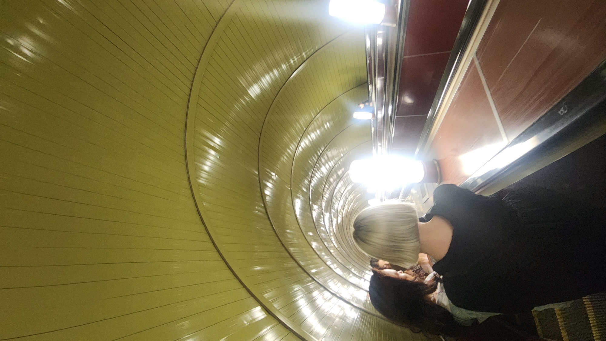 The image shows two individuals on an escalator inside what appears to be a tunnel-like structure. The tunnel has a yellow, ribbed interior. The escalator has a red side panel, with shiny lights overhead. The individuals appear to be a blonde-haired person in a black shirt and a dark-haired person behind them. The location is unknown.