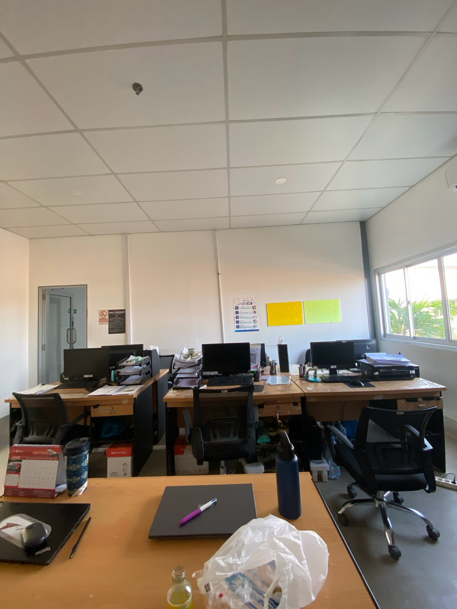 The image depicts a modern office space, likely in Batam, Indonesia, characterized by rows of workstations and a clean, well-lit environment. The ceiling features a grid of acoustic tiles with recessed lighting and a fire sprinkler head visible in the upper left quadrant. The walls are painted a neutral white, and a large window on the right side allows natural light to illuminate the room, offering a view of lush green palm trees and foliage, suggesting a tropical or subtropical setting.
The office is equipped with multiple computer workstations, each consisting of a desktop monitor, keyboard, and mouse. These are arranged on wooden desks, which are further organized into interconnected cubicle-like structures. Some desks have additional office supplies such as paper sorters, binders, and a printer. A black office chair with a mesh back is positioned at one of the desks in the foreground. On a nearby desk, a laptop, a stylus, and a bottle of what appears to be a beverage are visible. A crumpled plastic bag lies on the desk in the foreground, with a bottle of liquid next to it. A desk calendar from 2020 is also present, indicating a recent but not current time frame.
A doorway with a glass door and silver handles is visible on the left side, leading into another room or corridor. A sign with red and black lettering is affixed to the wall next to the door, possibly indicating office rules or a checklist. Two colored rectangles, one yellow and one lime green, are displayed on the wall further down, potentially for decorative or organizational purposes. The floor appears to be a neutral color, possibly tiled or linoleum. The overall impression is that of an organized and functional office environment, likely in a commercial or administrative building within Batam, Indonesia.