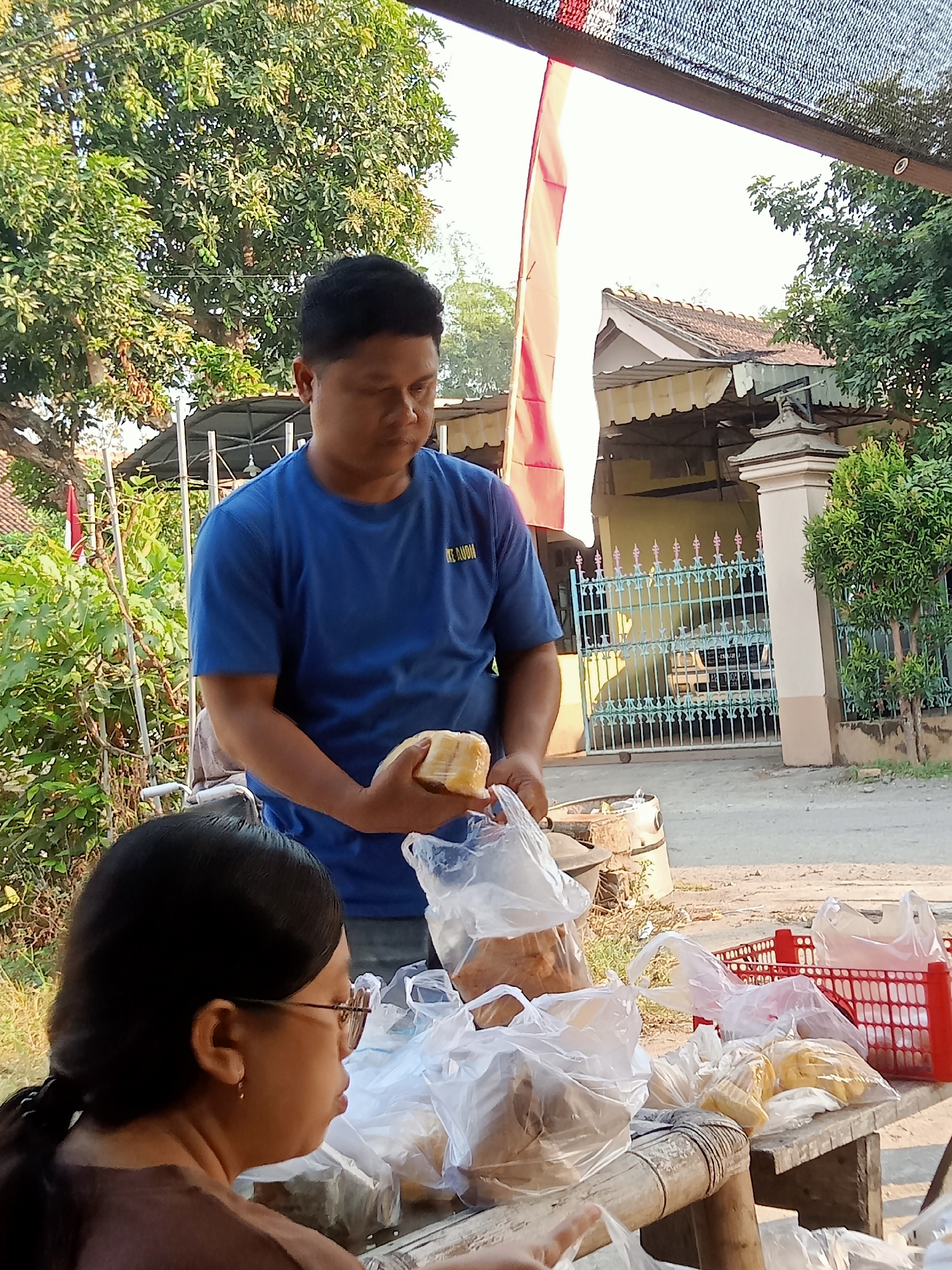 A man in a blue t-shirt is standing and holding a food item wrapped in plastic. He is in front of a table laden with more plastic-wrapped food items. To the left, a woman wearing glasses and a brown shirt is seated, facing the table. The scene appears to be outdoors, with trees, a building with a tiled roof, and a decorative metal fence in the background. The overall impression is of a food stall or a casual outdoor market setting. The presence of an Indonesian flag in the background suggests the location is in Indonesia, and the name "Simo" is a district within Boyolali Regency, Central Java, Indonesia, which fits the visual context of a residential area with a local market.