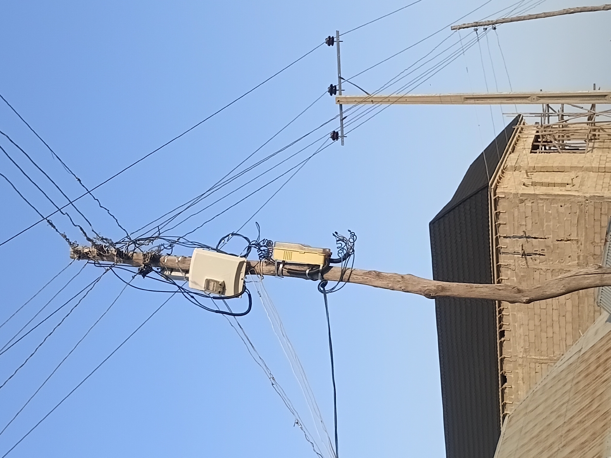 The image depicts an outdoor scene in Jalingo, Nigeria, under a clear blue sky. In the foreground, a weathered wooden utility pole stands vertically, supporting a complex network of electrical wires. Attached to the pole are two beige electrical enclosures, one above the other, with visible wiring and conduit. Below the utility pole, a building under construction is visible, with walls constructed from light-colored bricks. The building has a dark-colored, pitched roof. In the midground, another wooden utility pole with insulators and wires is visible to the right of the first pole. The environment appears to be an urban or semi-urban area, given the presence of utility infrastructure and construction.