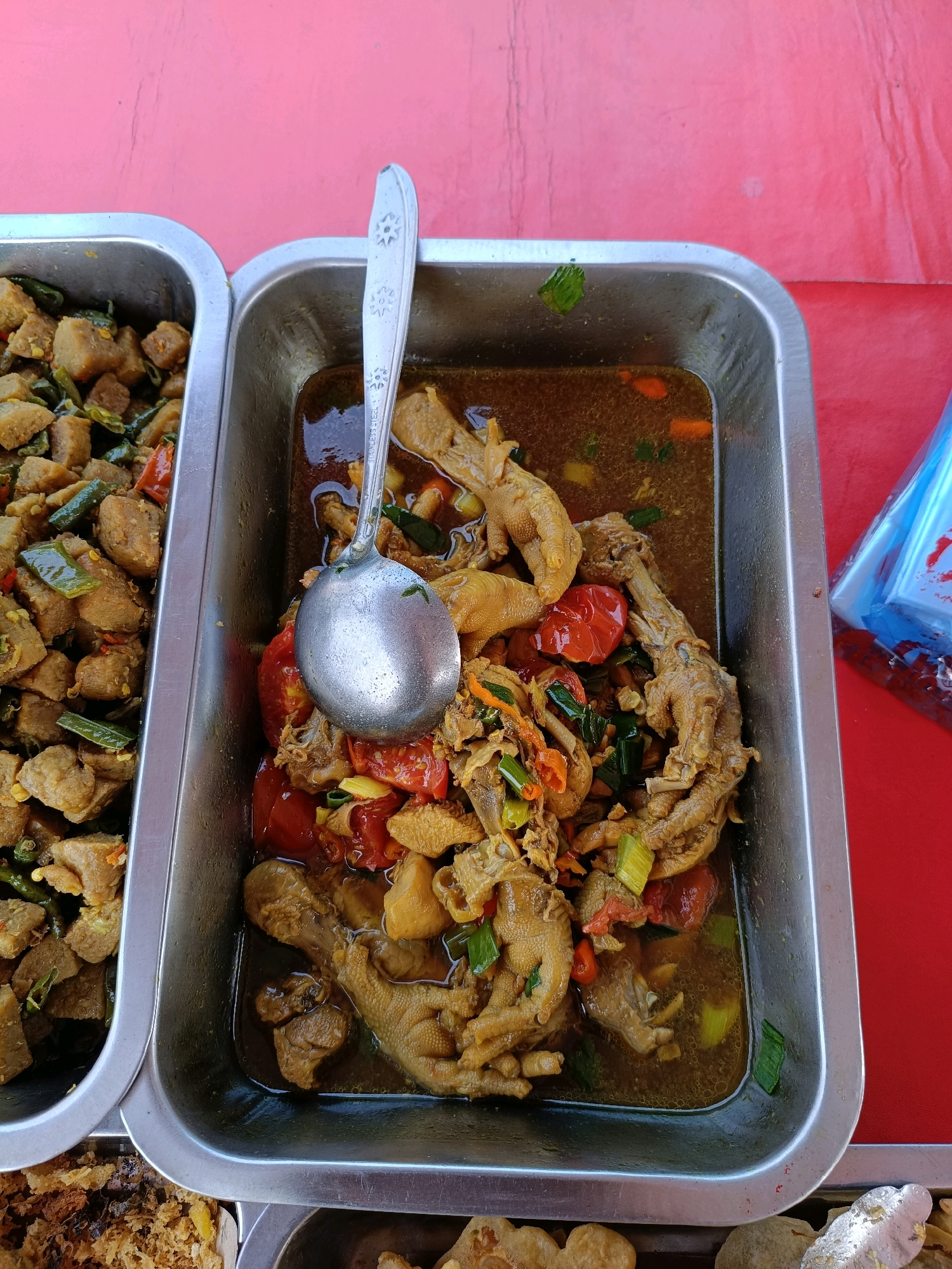 In Maguwoharjo, Indonesia, a metal tray filled with chicken feet stew is presented on a red surface. The stew consists of whole chicken feet, chopped tomatoes, green onions, and other ingredients in a dark, savory broth. A silver ladle rests on top of the food, its handle extending out of the frame. Adjacent to this tray, another metal tray contains a dish with cubed pieces of food, possibly tofu or meat, mixed with green beans and chili peppers. The overall scene suggests a food stall or market setting.