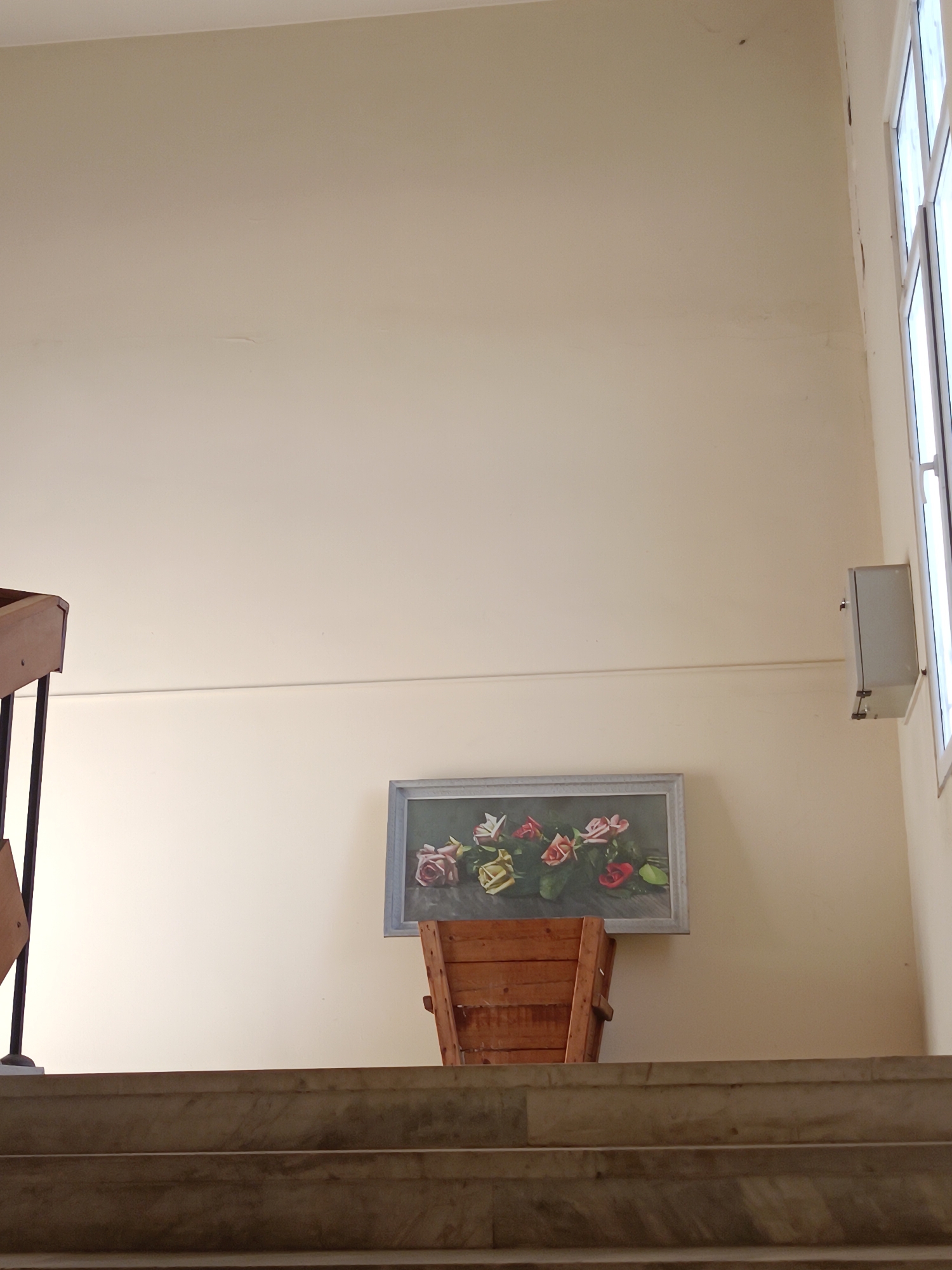 The image shows an interior scene, viewed from the bottom of a staircase. The wall is off-white and has a painting of flowers hanging on it. A wooden chest stands beneath the painting. To the right of the painting, there is a grey box mounted on the wall and a window letting in light. The location appears to be indoors. The location in the city of Plovdiv, Bulgaria is not discernible from the visual information.