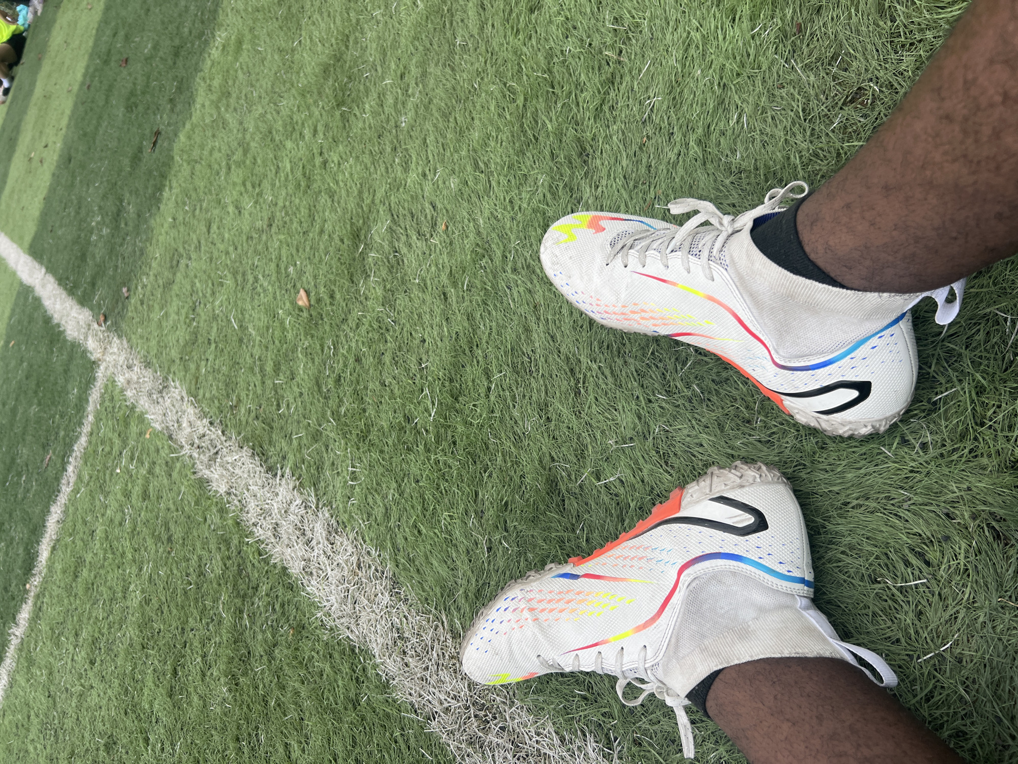 The image shows a view from above of a person's legs and feet wearing white soccer cleats with colorful accents, standing on a green artificial turf field. A white line, likely a field marking, is visible in the lower left of the frame. The person is wearing black socks. The image was not taken in Jiangjin, China.