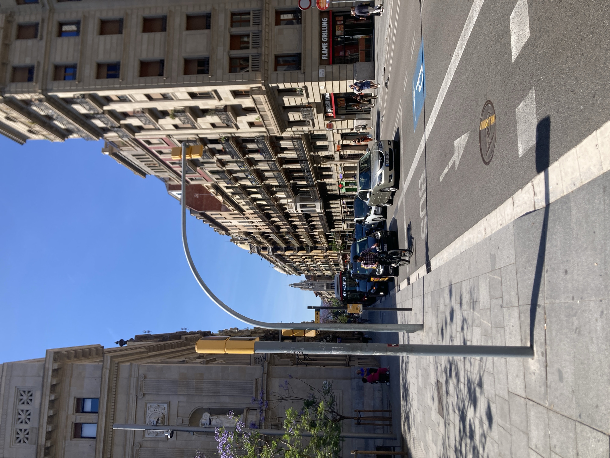 The image shows a street scene in Barcelona, Spain. The street is lined with tall, multi-story buildings with balconies. A Burger King "Flame Grilling" restaurant is visible on the right side of the street. There are vehicles parked along the street, including cars and a motorcycle. A city bus is also visible. People are walking on the sidewalks and crossing the street. Streetlights and signs are present. The sky is clear and blue. There is a tree with purple flowers in the foreground. The photo is taken at an angle, with the perspective skewed.