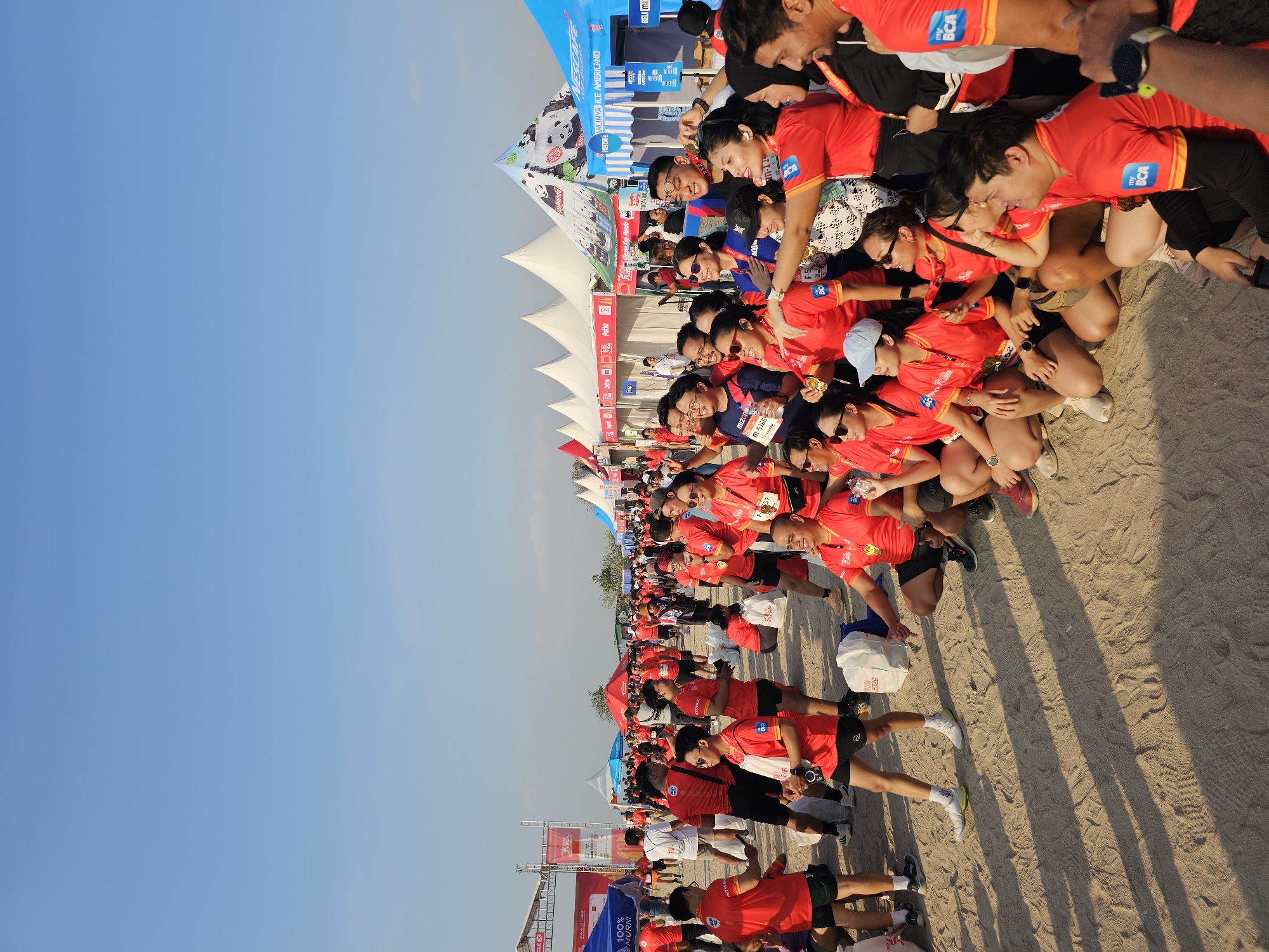 A group of people, mostly wearing red shirts with "my BCA" logos and some with race bibs, are gathered on a sandy surface, likely a beach or park in Denpasar, Indonesia. The sky is clear and blue, indicating daytime with bright sunlight casting shadows on the sand. In the background, there are several tents set up, some with event branding, suggesting an outdoor event like a marathon or festival. Some individuals are posing for photos, while others are interacting and standing around bags that appear to contain event merchandise. The overall atmosphere seems to be celebratory and social.