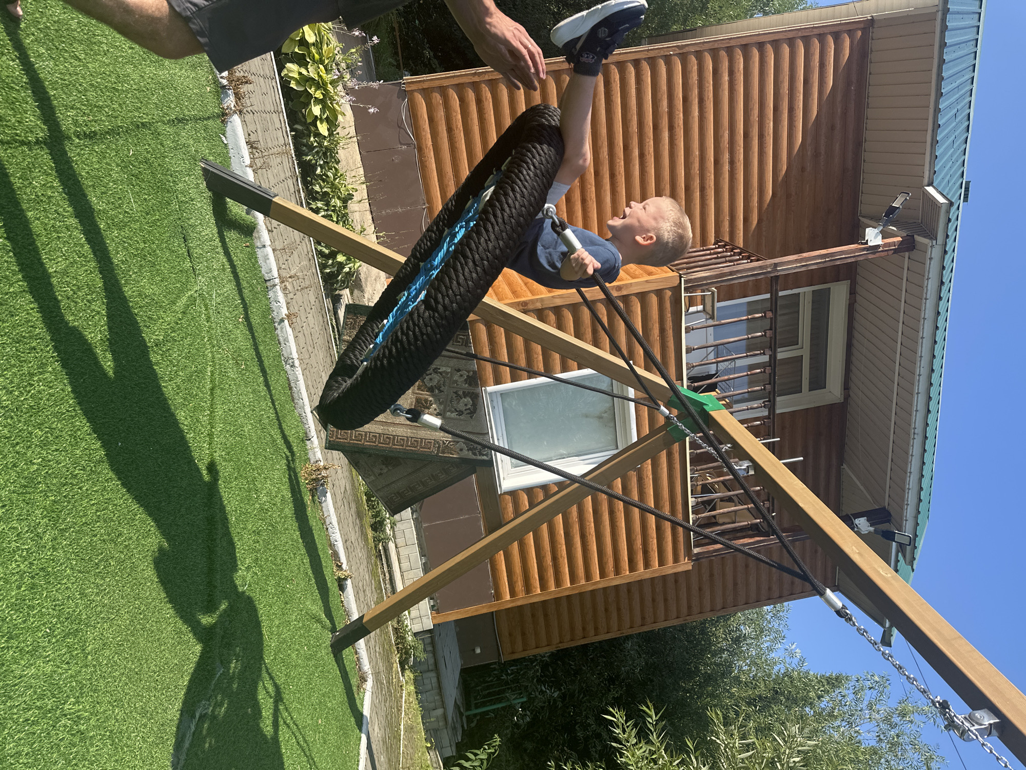 A young, blond boy with fair skin is joyfully swinging on a tire swing in a backyard. He is wearing a dark blue t-shirt and grey shorts, with one leg extended forward and the other bent. His mouth is open in a wide smile or laugh. His right hand is holding onto a grey handle attached to the swing. The swing itself is made from a black tire and has blue rope attached. A person's arm, likely an adult, is visible on the left side of the frame, reaching out towards the boy, possibly assisting with the swing.
The background features a rustic, wooden house with a green metal roof. The house has visible log construction and a small balcony with a wooden railing. There is a window on the side of the house. The yard is covered with artificial turf, and a swing set made of wooden beams supports the tire swing. The overall environment suggests a sunny day with clear blue skies and shadows cast by the swing set and trees. The location is identified as Danilovka, Altai Krai, Russia.