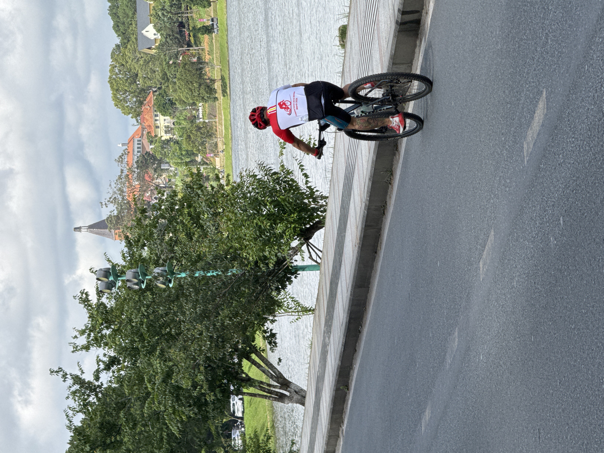 A person wearing a red helmet and a cycling jersey with red and white accents is riding a bicycle on a paved road. The cyclist is facing away from the camera, with their left arm resting on the handlebars and their right hand also on the handlebars. They are wearing red and white cycling shoes and black shorts with teal accents. The bicycle has a water bottle cage on the frame. The road has white dashed lines marking lanes. To the left of the road is a raised concrete sidewalk with textured gray panels. Beyond the sidewalk is a body of water, possibly a lake, with ripples on the surface. Trees with green foliage are growing along the edge of the water. In the background, there are buildings with red and orange roofs, some with white facades, nestled amongst more trees on a gentle slope. A tall, dark-colored tower with a pointed roof is visible in the distance. Further in the background, the sky is overcast with gray clouds. There is a traffic light pole with multiple signal lights on the left side of the image, partially obscured by trees. The scene appears to be a scenic area in Phường Xuân Hương, Đà Lạt, Vietnam, likely near a lake or park.