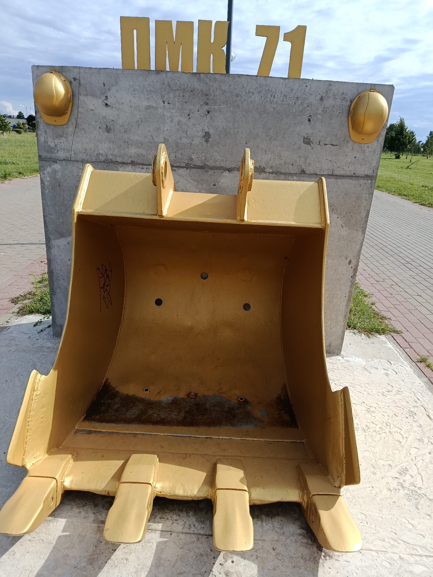 The image depicts a large, golden excavator bucket standing upright on a concrete base. Above the bucket, a concrete slab bears the Cyrillic letters "ПМК 71" in large, golden block letters. Two golden hard hats are mounted on the sides of the concrete slab. The scene appears to be an outdoor public space, possibly a park or memorial, with a paved walkway and grass visible in the background under a cloudy sky. The excavator bucket has a graffito tag on its interior surface. The monument is located in Zhlobin, Belarus.