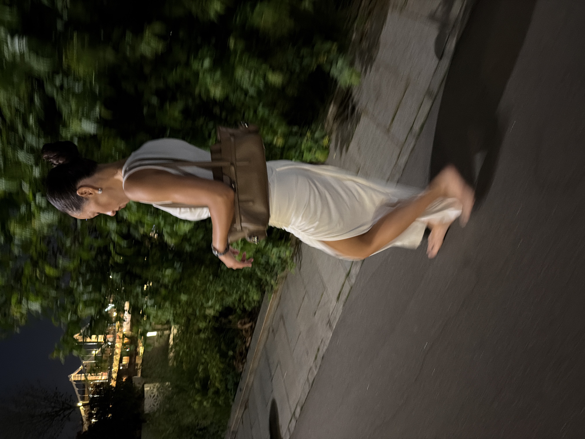A woman walks barefoot on a paved path at night, her motion creating a blur. She wears a flowing white dress with a high slit and carries a brown handbag. Behind her, lush green foliage lines the path, and in the distance, a bridge with illuminated structures is visible. The ground is dark asphalt with some scattered white flecks, and the path is made of light-colored concrete slabs. The overall scene suggests an urban environment at night, possibly near a body of water, with an architectural landmark in the background.