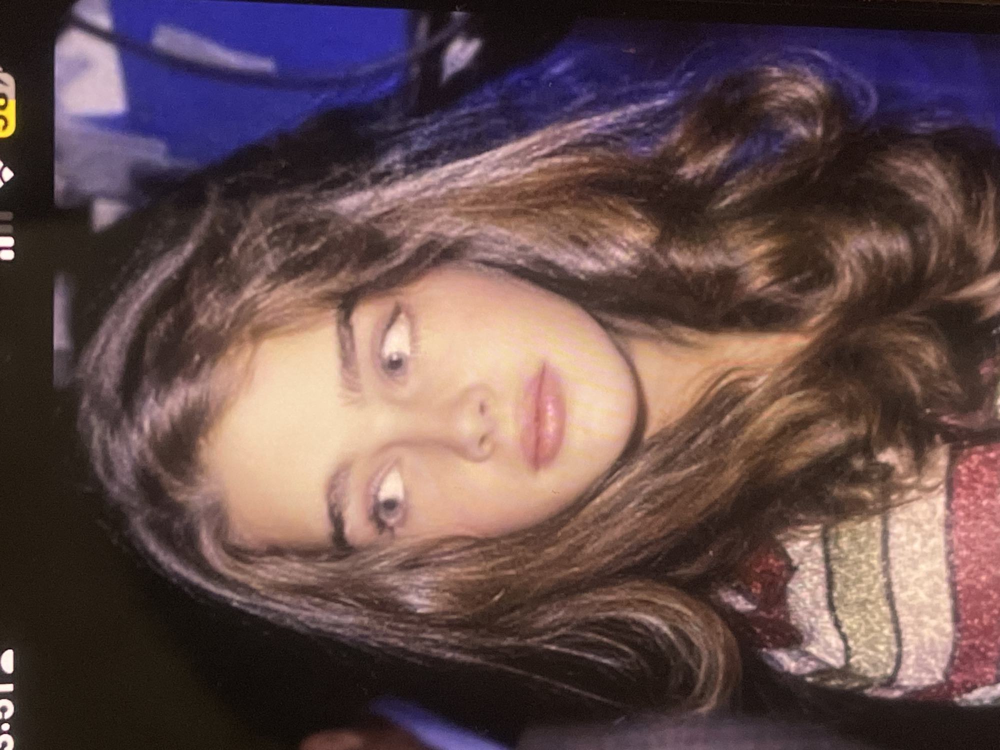 The image displays a close-up portrait of a young female, positioned at an angle, rotated clockwise. She has long, wavy, voluminous brown hair, fair skin, full pink lips, and prominent dark eyebrows. Her large, light-colored eyes are wide open and looking upwards and slightly to the left, conveying an expression of intensity, surprise, or wonder.
She is wearing a top or garment with vertical stripes in shades of red, white, and a muted green or gold, which appears to have a glittery or textured finish. The background behind her is a solid, deep blue, suggesting an indoor setting, possibly a studio with a colored backdrop.
The photograph itself appears to be an image of a screen, indicated by the visible dark bezel on the left and top edges. On the left side of the screen, elements of a phone's user interface are partially visible, including a battery icon with a yellow fill, likely indicating a charge percentage (e.g., "50%"), and a signal strength indicator. The digital time "5:51" is partially visible at the bottom left of the screen.
No specific visual cues in the image itself denote the location context of Poltava, Ukraine, as it is a generic portrait. The overall scene is a brightly lit, posed shot of the subject.