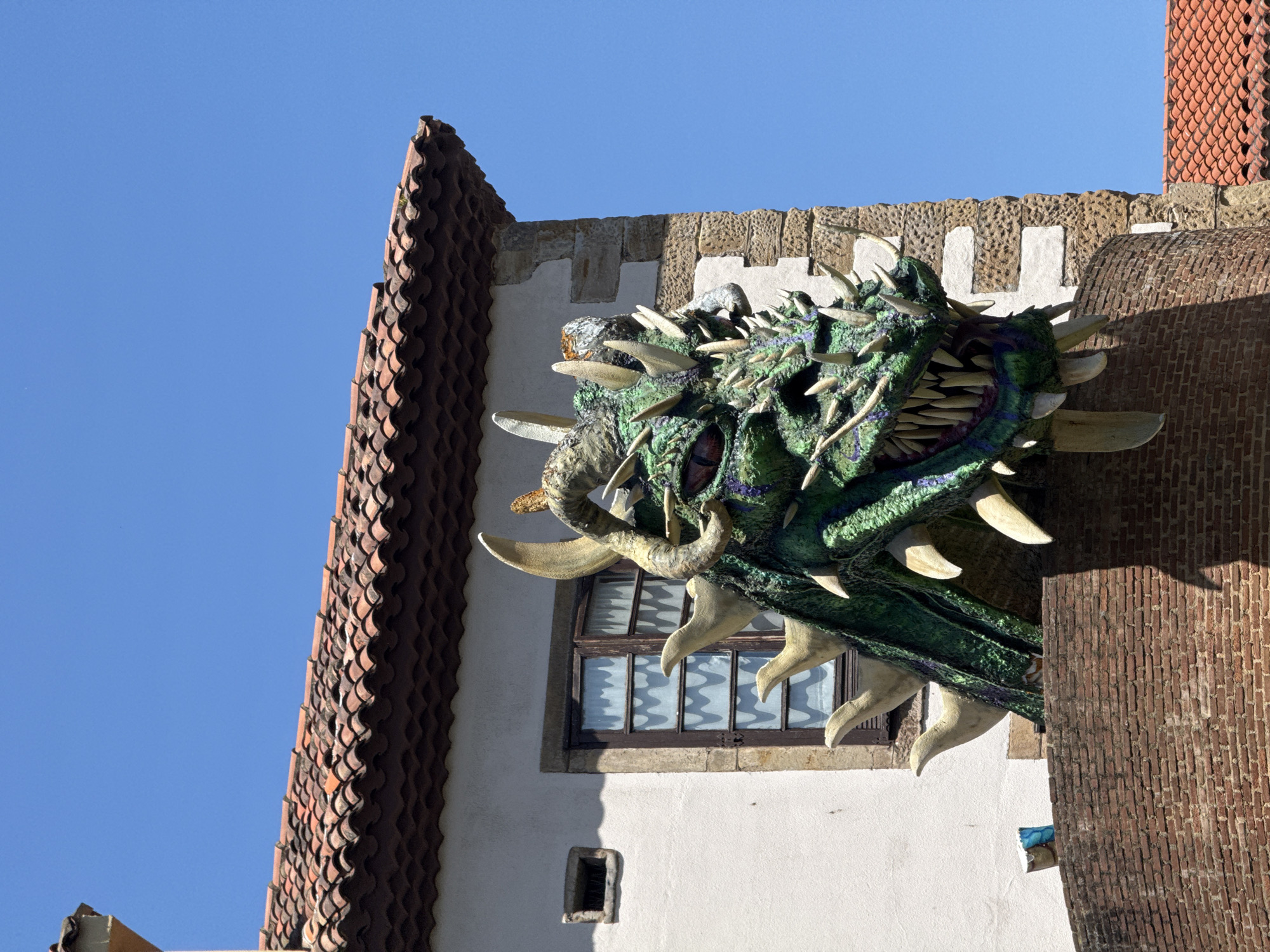 A green dragon sculpture with spines and horns is attached to a building in Gijón/Xixón, Spain. The dragon's head and part of its neck are visible, protruding from the corner of the building. The building has a combination of architectural styles, including a white stucco wall with a window, a brick facade, and a roof covered in red clay tiles. The sky above is clear and blue.