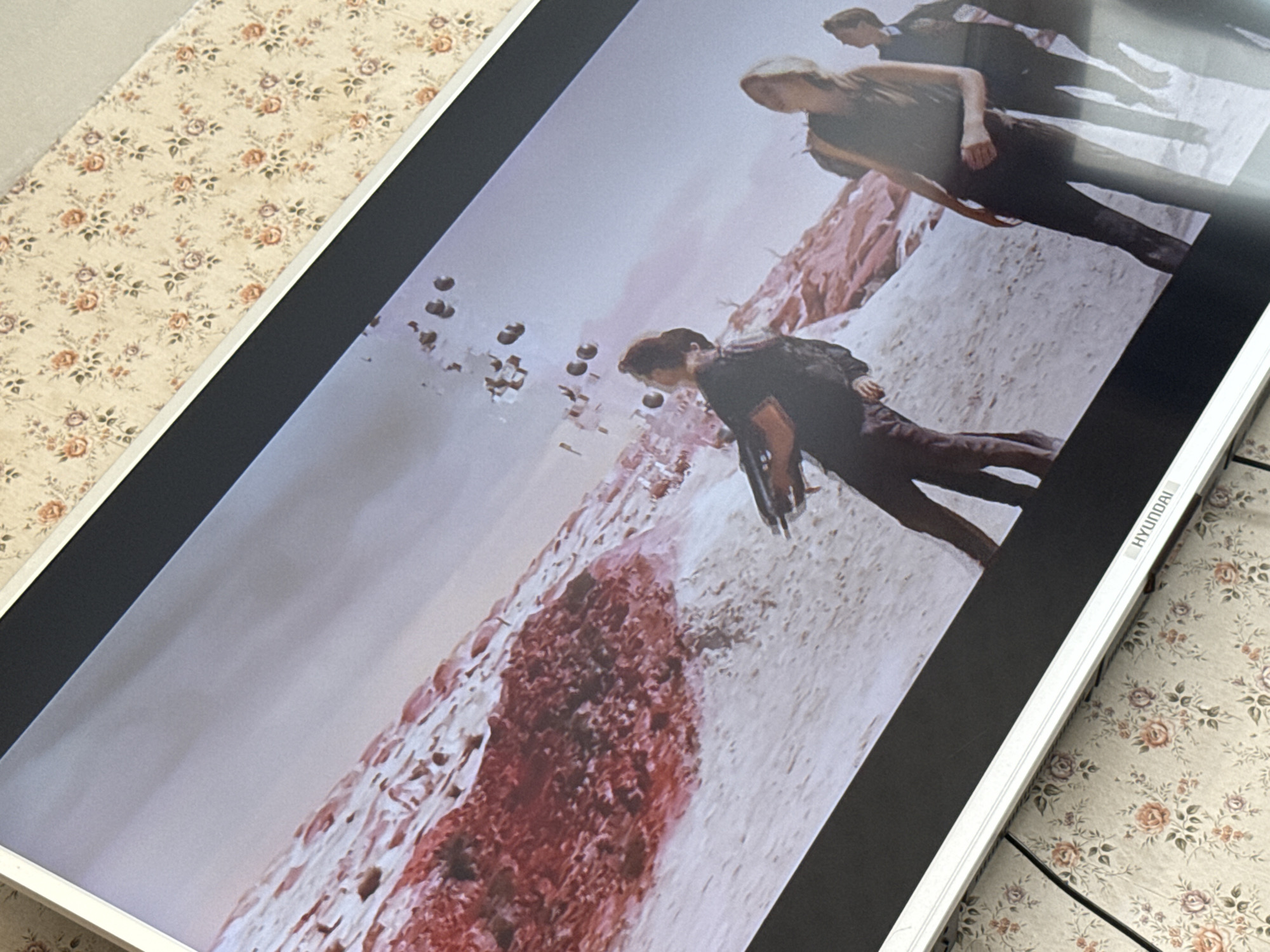 The image shows a Hyundai TV screen displaying a scene with several individuals in a desolate, otherworldly environment. Three figures are visible, two in the upper portion of the screen walking away from the camera at an angle, and a third closer to the center-left of the screen. The landscape appears to be a barren, rocky terrain with red-tinged formations. Numerous small, dark objects are scattered in the sky. The scene appears to be from a science fiction or fantasy movie or show. The TV is sitting on a surface with a floral pattern. The location is Mytishchi, Russia, cannot be determined from the image.