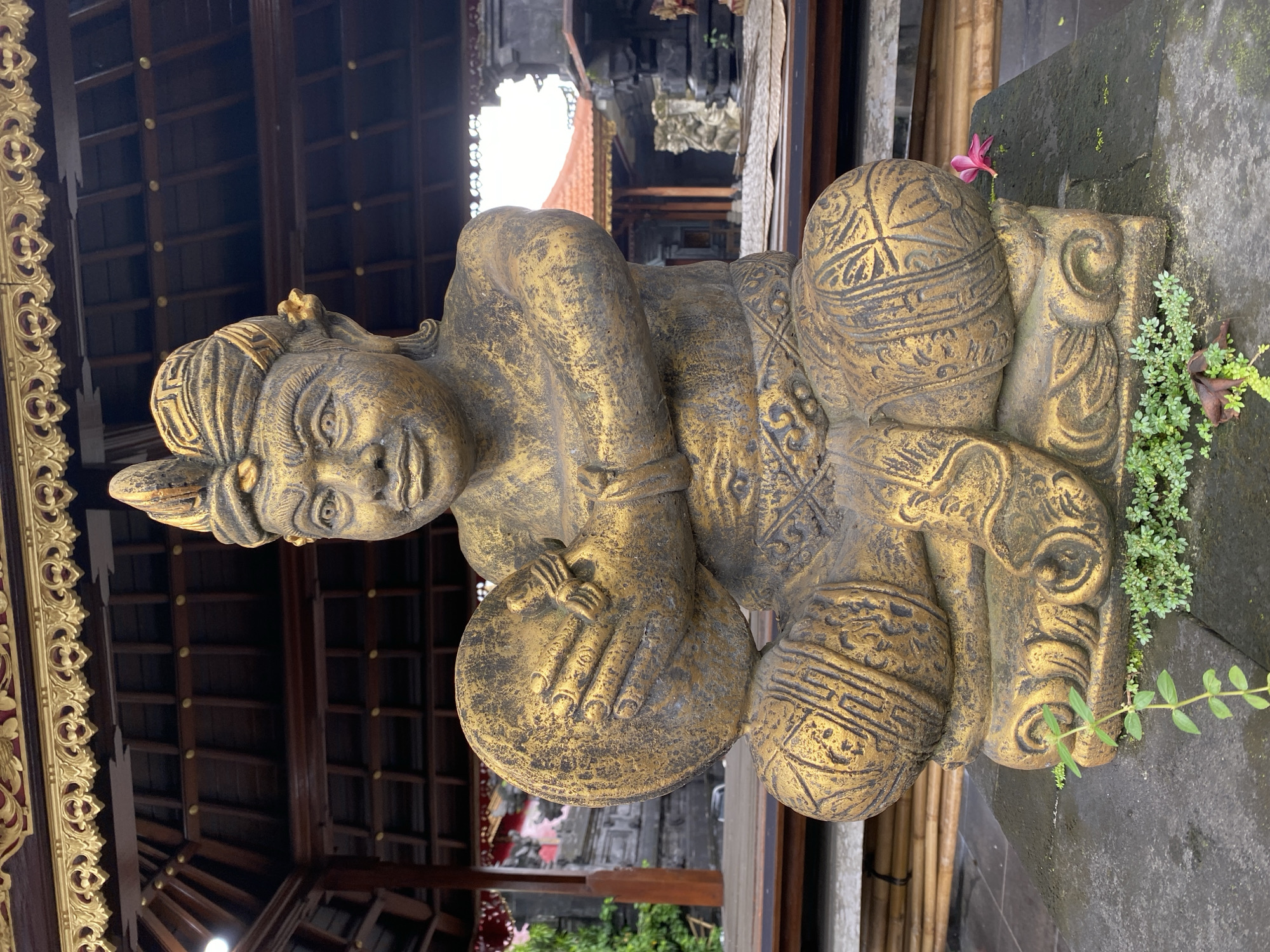 The media file displays a weathered golden stone statue, likely depicting a male figure, situated within a traditional Balinese architectural setting in Kuta Selatan, Indonesia. The statue is shown in a seated or reclining pose, facing towards the viewer with a serene expression. It features an elaborate, patterned headpiece and intricate carvings on its body and attire, hinting at traditional clothing or ceremonial regalia. The figure's hands rest on or hold two spherical, pot-like objects, also adorned with patterns. The golden paint on the statue is aged, with darker patinas and areas where the color has faded, revealing the stone beneath.
Immediately surrounding the statue, small green plants and moss cling to the stone base and the adjacent ground, with a delicate pink flower blooming close to the statue's back. In the background, dark wooden ceilings with visible beams and rafters create a structured canopy. To the left, ornate golden carvings embellish a wooden frame or pillar, reflecting the rich decorative style characteristic of Balinese architecture. Further in the distance, glimpses of other traditional buildings and a bright sky are visible.
The scene appears to be an outdoor area of a temple, palace, or cultural compound, typical of Indonesia. The lighting suggests it is daytime, with soft, indirect illumination. No specific activity or event is taking place; the image captures a static cultural artifact within its historical context. There is no visible text in the image.