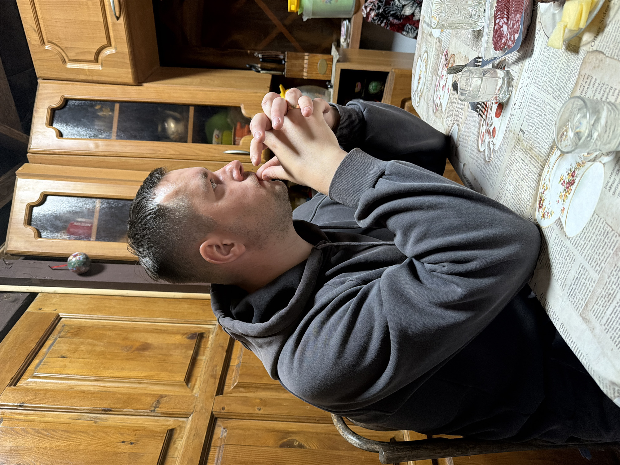 A Caucasian man with short dark hair and stubble is seated at a table, wearing a dark grey hooded sweatshirt. He is looking to his right, with his hands clasped in front of his mouth. The table is covered with a tablecloth printed with newspaper-style text and images of teacups. On the table, there are several glasses, some containing clear liquid, a small plate with sliced salami, and a small dish with cheese. Behind the man, a wooden kitchen cabinet with glass-fronted doors is visible. The overall environment appears to be a home interior. The image was taken in the city of Krasny Mayak, Russia.