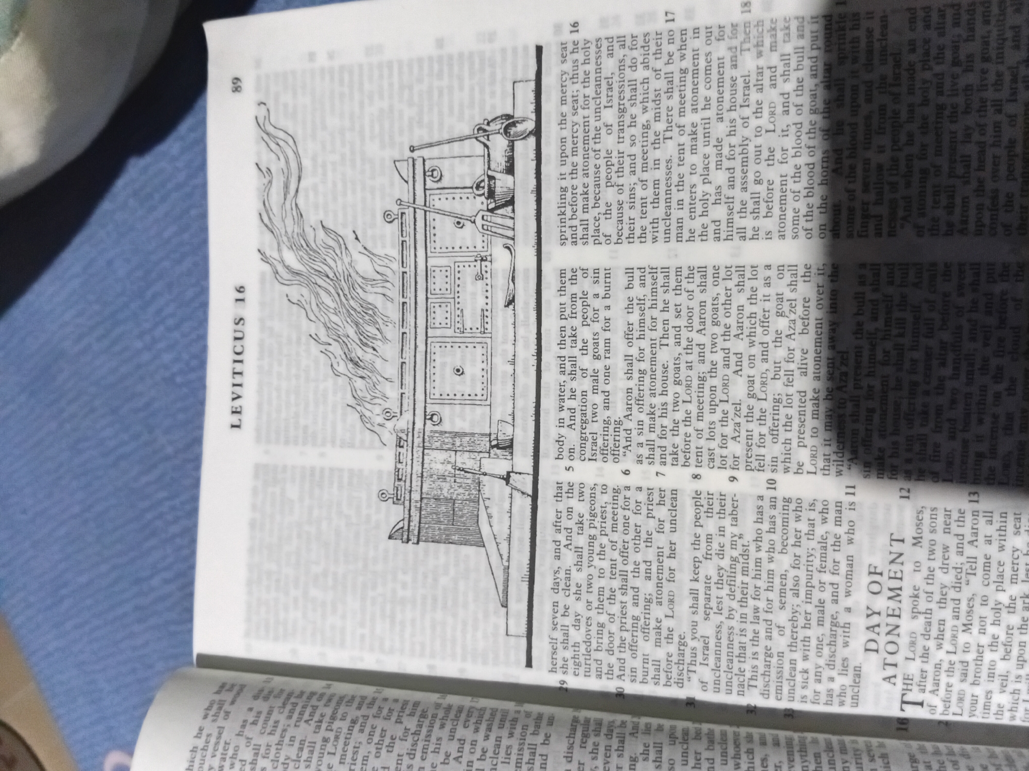 The image shows an open page of a book, likely a Bible, with text and an illustration. The text is in columns, with the book title "LEVITICUS 16" visible at the top, along with the page number "89". The illustration depicts a structure resembling the Ark of the Covenant, with flames rising from it, and various tools nearby. The page also contains headings and numbered verses of text. The book is lying on a blue fabric surface, possibly a tablecloth or blanket. The lighting suggests it is indoors, with shadows cast on the page. There are no people or animals visible in the image, and no indication of a specific city like Nkpor, Nigeria. The scene is static and focuses solely on the book and its contents.