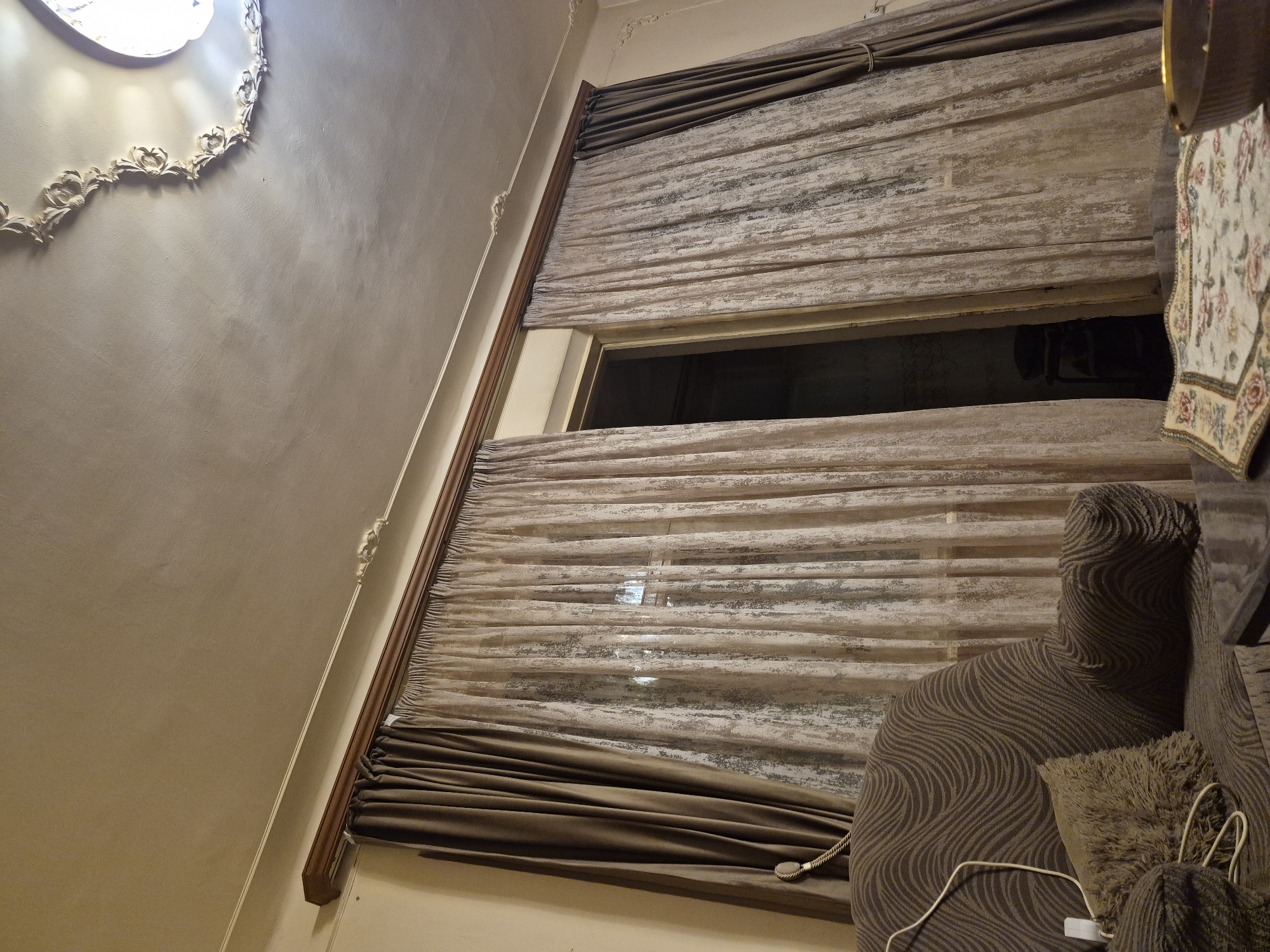 This interior image, stated to be in Yerevan, Armenia, depicts a dimly lit room, likely a living area, focusing on a window or doorway dressed with curtains and a section of a couch.
In the foreground, on the left, a portion of a brown upholstered couch covered with a textured slipcover featuring a wavy pattern is visible. A fluffy brown throw pillow rests on the couch, alongside a white electrical cable connected to a small white power adapter or strip. To the right, a dark, reflective surface (likely a table) supports a decorative fabric mat with a floral pattern in hues of pink, green, yellow, and blue on a cream background. A dark brown bowl with a gold rim is partially visible on the table.
The midground is dominated by a two-layered curtain assembly. The inner layer consists of sheer, light brown curtains characterized by a distressed or abstract vertical blotchy pattern. The outer layer comprises opaque, solid brown/taupe curtains, which are currently pulled back and secured with decorative rope tie-backs. These curtains hang from a dark wooden curtain rod, concealed by a matching wooden valance or fascia. A narrow vertical gap between the central sheer curtains reveals a dark, indistinct interior space beyond, where a multi-tiered shelving unit is faintly discernible.
The background features a light-colored ceiling with subtle texture and some discoloration. A white decorative molding runs along the upper perimeter of the wall. Sections of ornate white plasterwork are integrated into the molding and, more prominently, form a partial ceiling medallion surrounding a brightly illuminated light fixture located in the upper right quadrant of the image. The primary light source appears to be the ceiling fixture, casting a soft, artificial glow over the scene. No individuals are present.