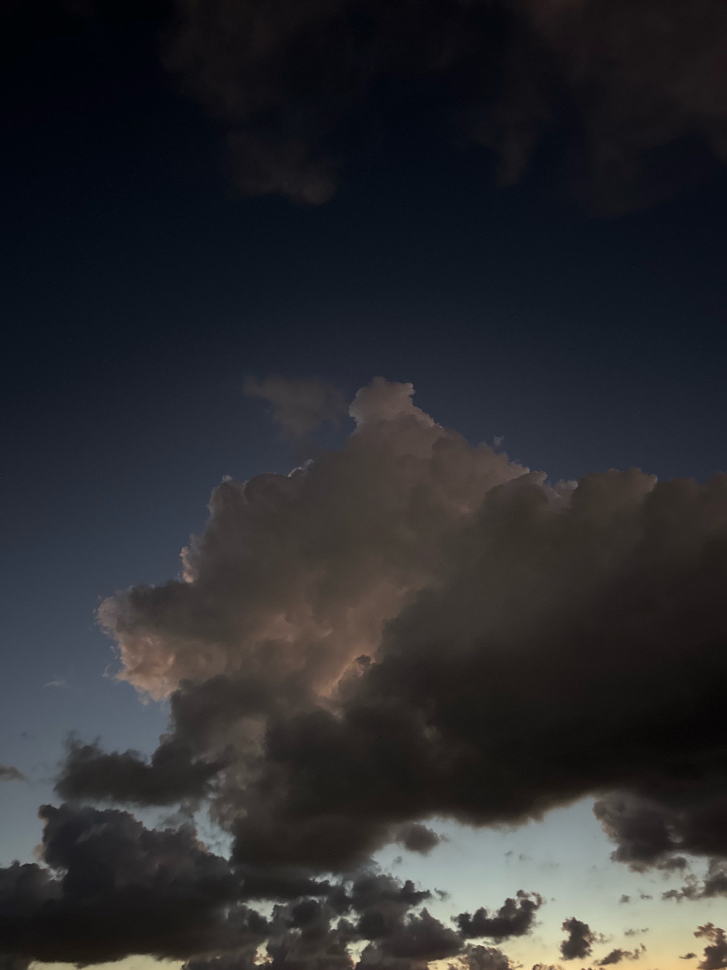 The image depicts a twilight sky with dramatic cloud formations over the city of Isola delle Femmine, Italy. The sky transitions from a deep, dark blue at the top to lighter shades of blue and hints of yellow near the horizon. A large, voluminous cumulonimbus cloud dominates the center of the frame, its edges illuminated by the last rays of sunlight, casting a warm, peachy glow on its underside. The cloud appears dark and imposing, with textured surfaces and clearly defined edges, suggesting dynamic atmospheric conditions. Smaller, fragmented clouds are scattered throughout the lower portion of the image, some appearing as dark silhouettes against the lighter sky. The overall impression is one of a moody and atmospheric sunset over a coastal Italian landscape.