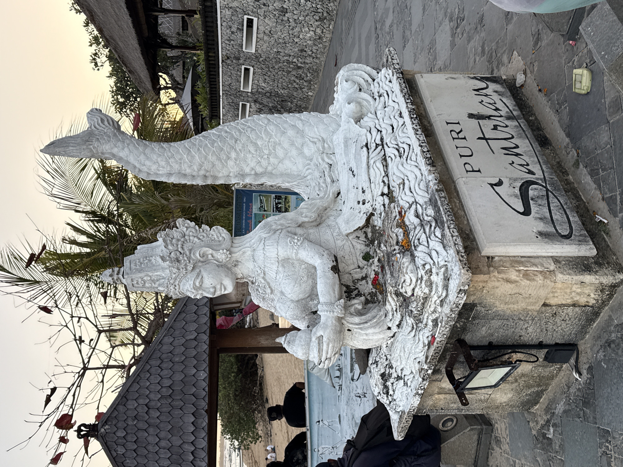 The image shows a white stone statue of a mermaid-like figure in a seated position, with a fishtail and elaborate headdress. The statue is situated on a stone pedestal with weathered lettering that reads "PURI Santrian". The pedestal is surrounded by a decorative stone base with wave-like carvings. Scattered petals of red and yellow flowers are visible on the pedestal and around the base of the statue.
To the left of the statue, there is a small, weathered wooden gazebo with a dark, hexagonal-patterned roof. In the background, behind the gazebo, a sandy beach is visible with several people sitting and standing. The sky is a pale orange, suggesting it is either sunrise or sunset. Palm trees and other vegetation are present in the background, as well as thatched-roof structures, indicative of a tropical resort or coastal area.
A metal spotlight is mounted on the side of the pedestal, facing outwards. A blue sign with text and images is partially visible behind the statue. The ground is paved with stone tiles. A light-colored backpack is on the ground to the left of the pedestal. A small, light green object, possibly a box or container, is on the ground in the lower right corner of the image.
Given the lettering "PURI Santrian" on the pedestal, this statue is likely located at Puri Santrian Hotel in Sanur, Denpasar, Bali, Indonesia. Sanur is a coastal resort town in Denpasar, known for its beaches and hotels.